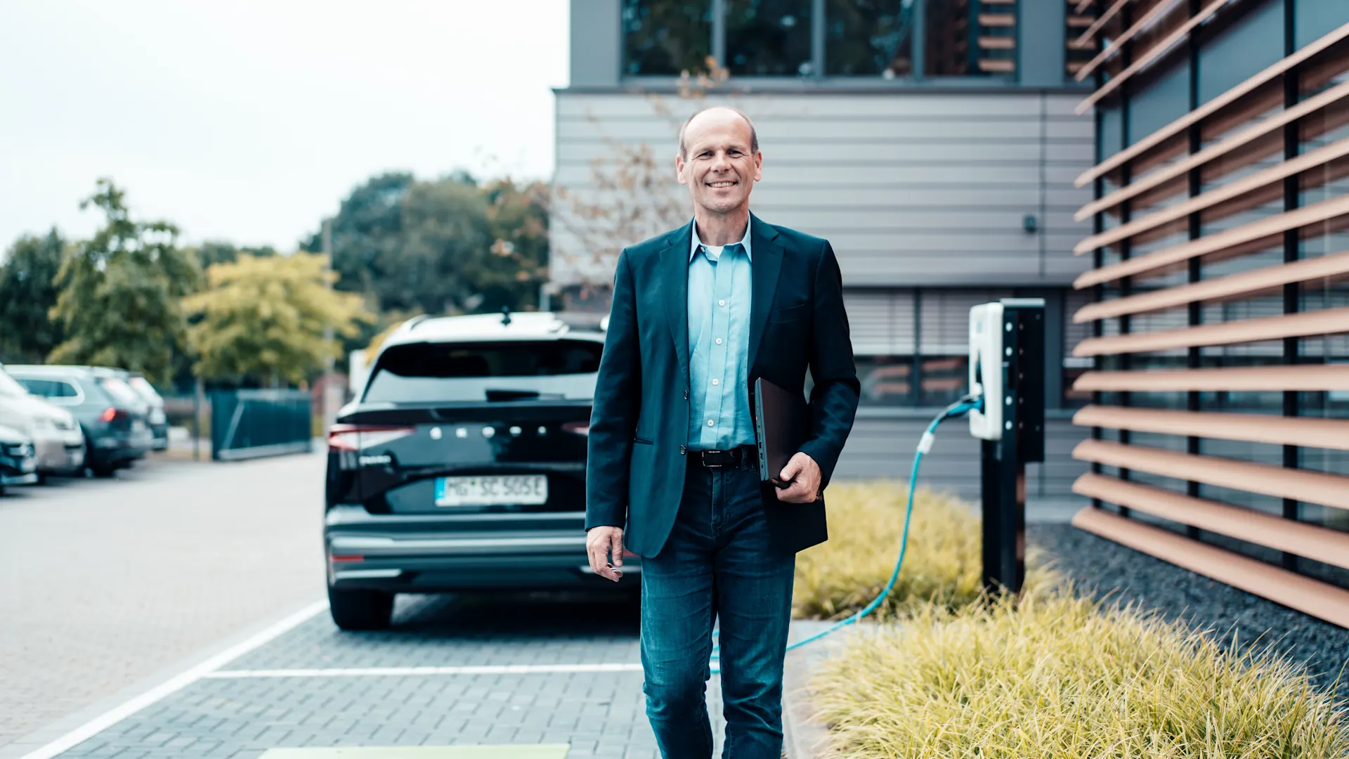 Unternehmer lädt mit einer Wallbox das Auto Unternehmer geht von seinem E-Auto weg und hält einen Laptop in der Hand, was mithilfe einer Wallbox auflädt.