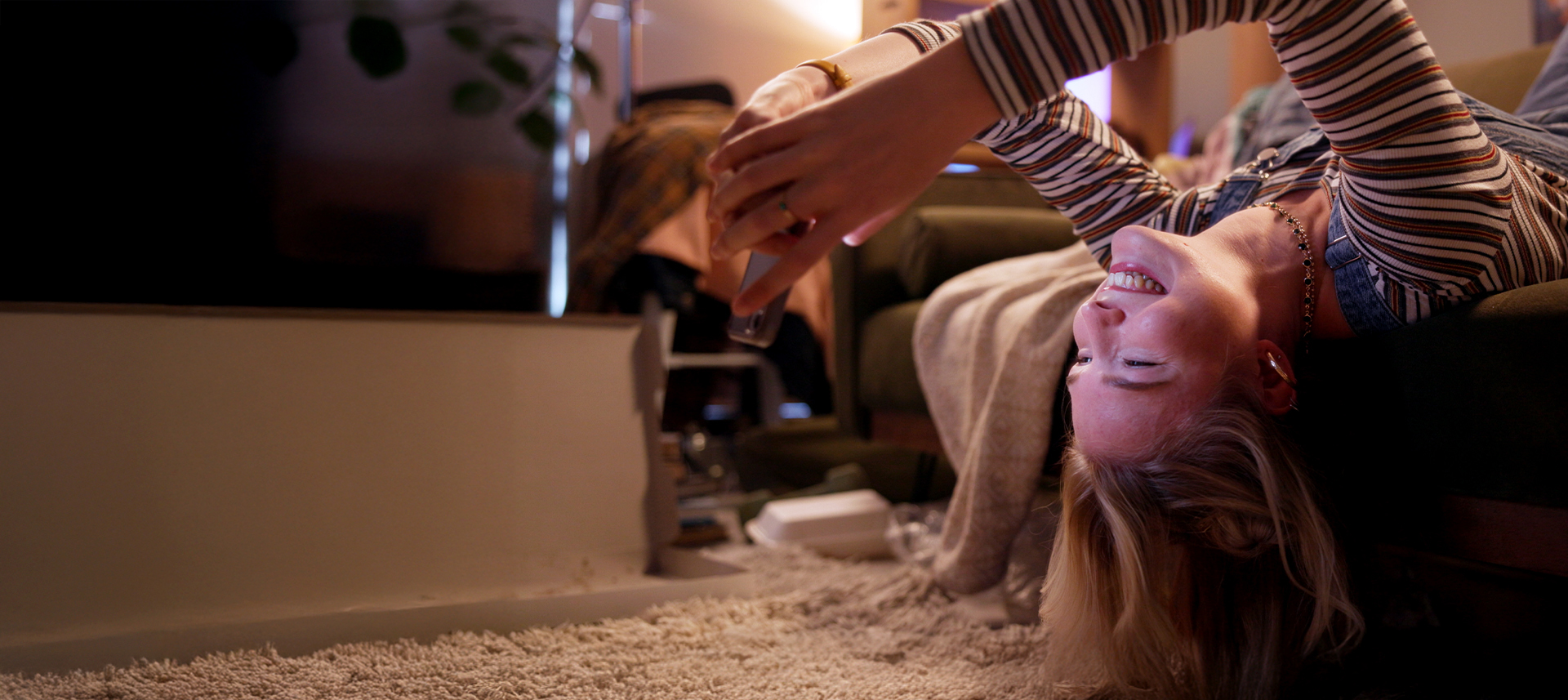 Frau liegt kopfüber von ihrem Sofa und schaut lachend auf ihr Smartphone. 