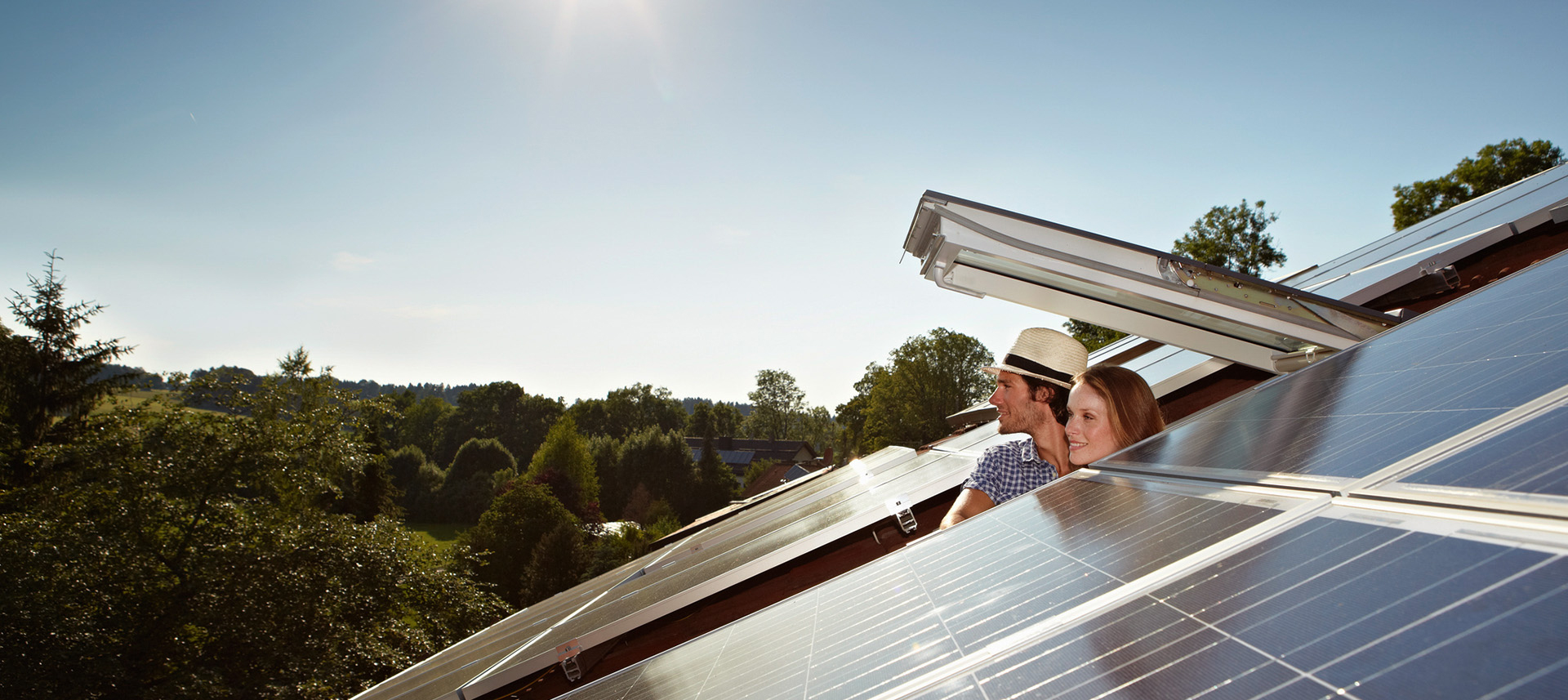 Mann und Frau schauen aus einem geöffneten Dachfenster. Eine PV-Anlage der NEW Energie ist zu sehen.