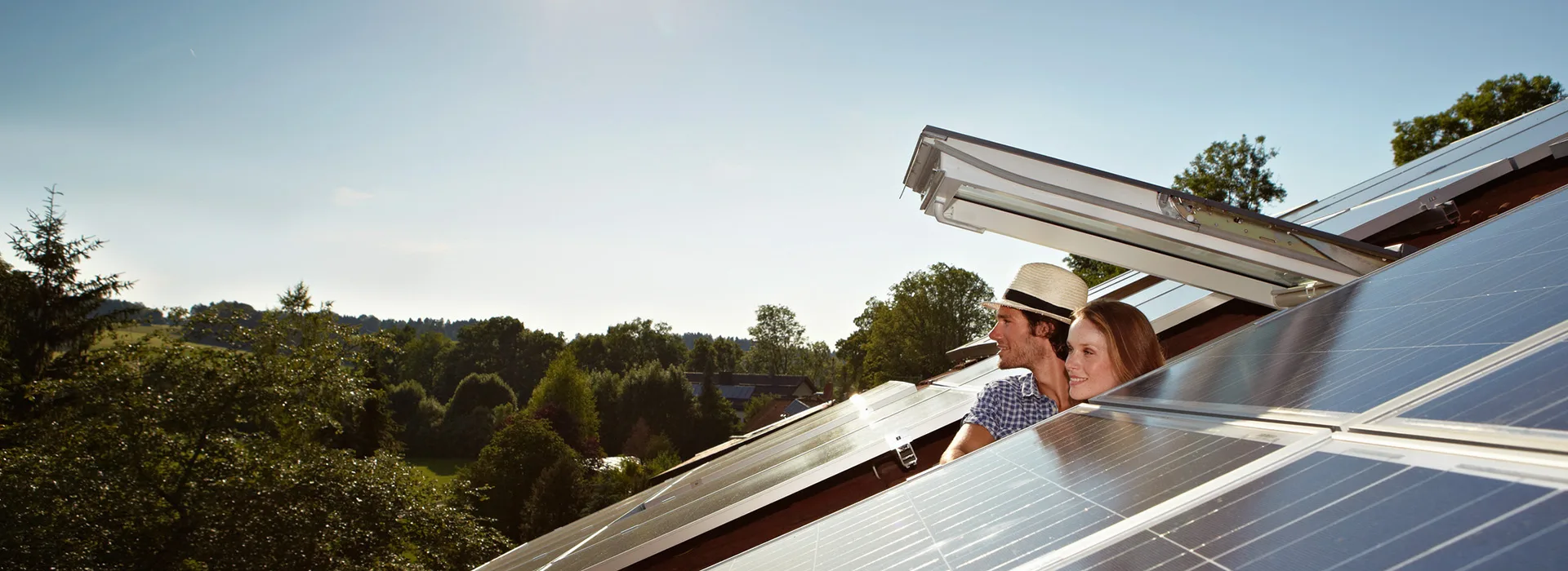 Mann und Frau schauen aus einem Dachfenster in die Ferne Mann und Frau schauen aus einem geöffneten Dachfenster. Eine PV-Anlage der NEW Energie ist zu sehen.