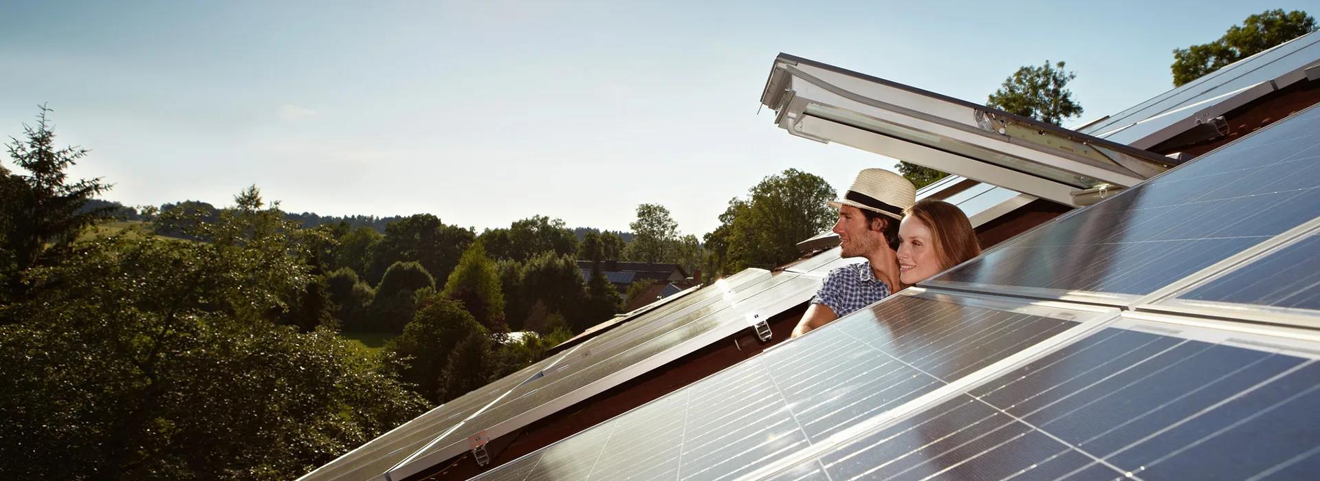 Mann und Frau schauen aus einem Dachfenster in die Ferne Mann und Frau schauen aus einem geöffneten Dachfenster. Eine PV-Anlage der NEW Energie ist zu sehen.