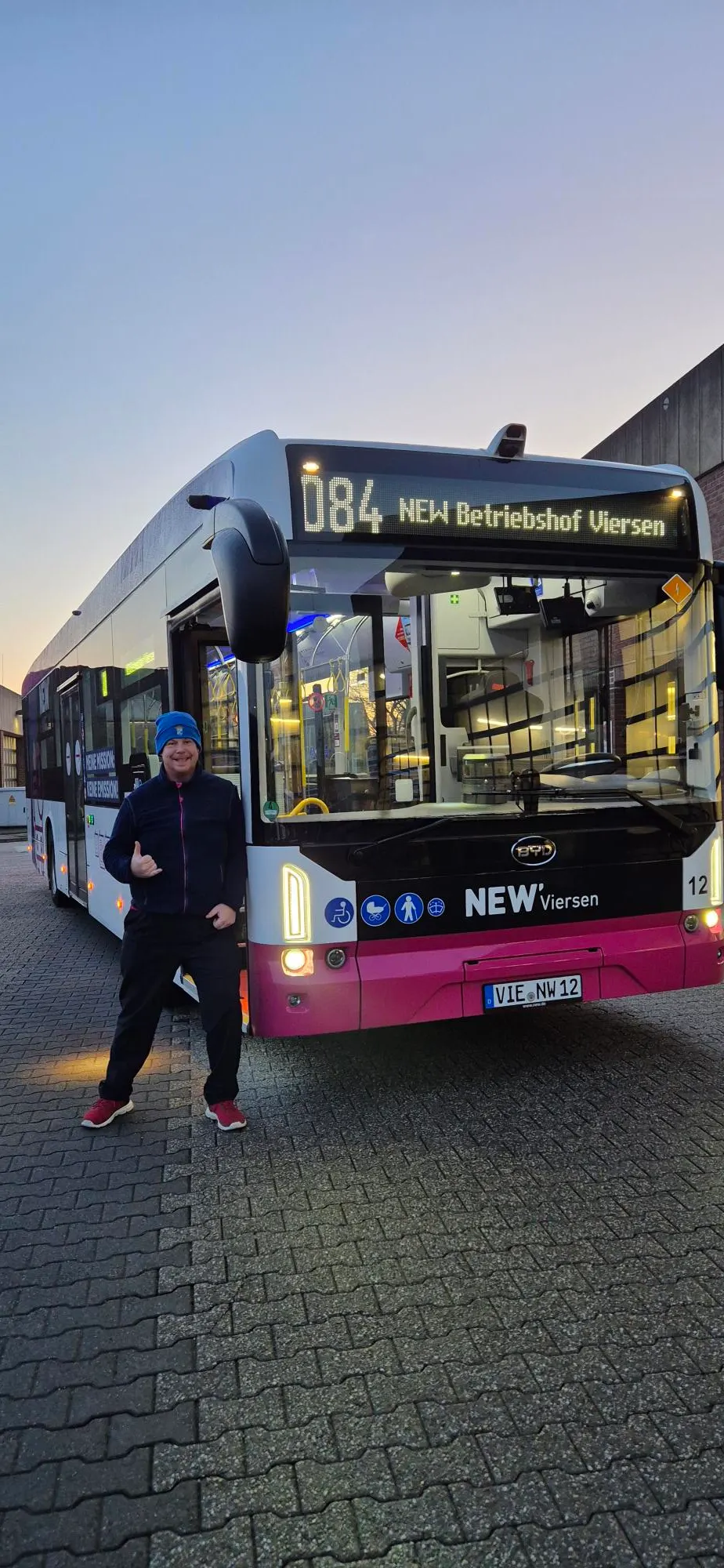 Martin Giesers-Aviles in Sportkleidung und blauer Mütze steht lächelnd mit Daumen hoch vor einem weißen Elektrobus der Linie 084 mit der Digitalanzeige NEW Betriebhof Viersen in der Abenddämmerung.
