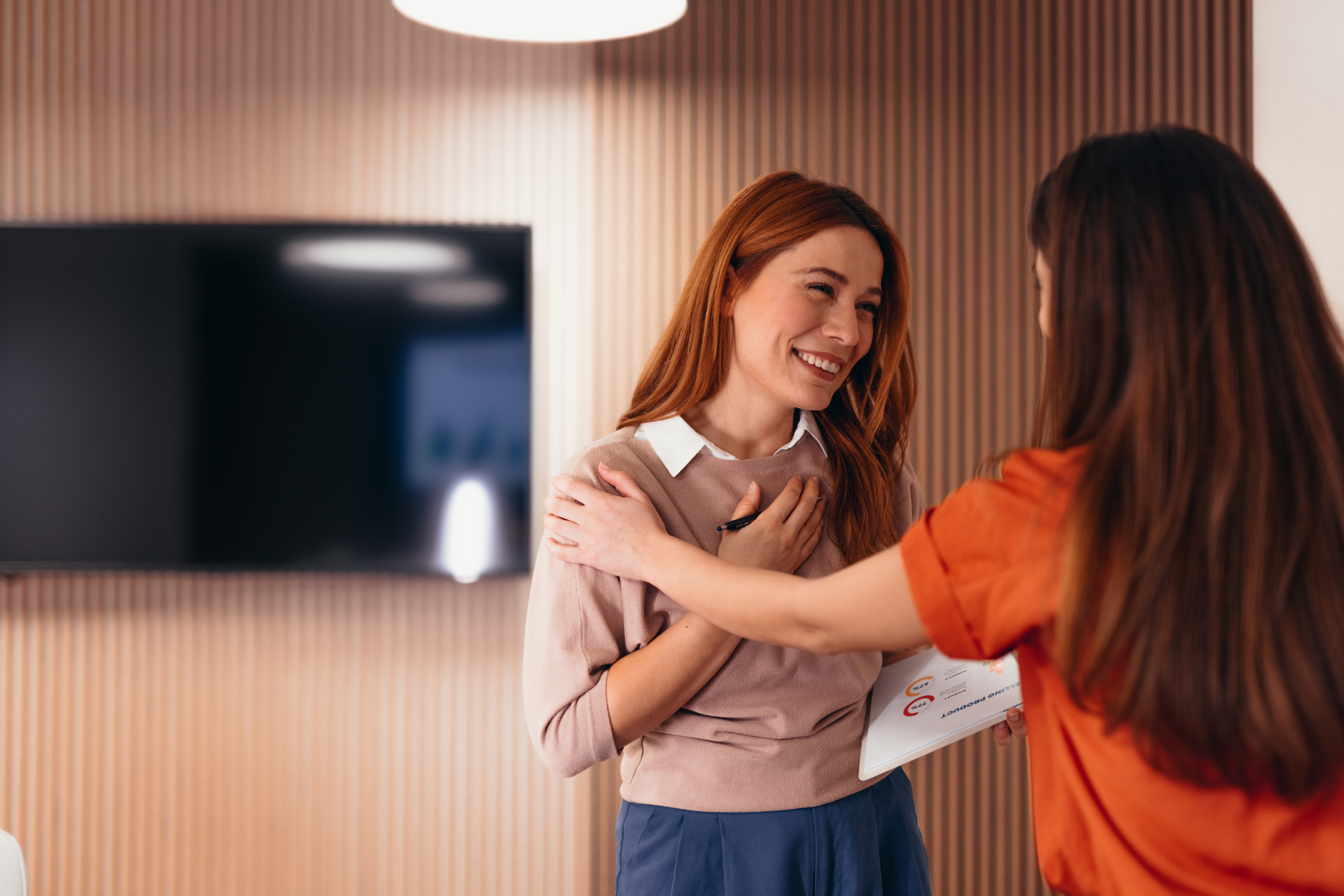 Zwei Frauen begrüßen sich herzlich in einem modern eingerichteten Raum, wobei eine rothaarige Frau in beigem Oberteil lächelnd ihre Gesprächspartnerin in orangefarbener Kleidung die Hand auf die Schulter legt. 