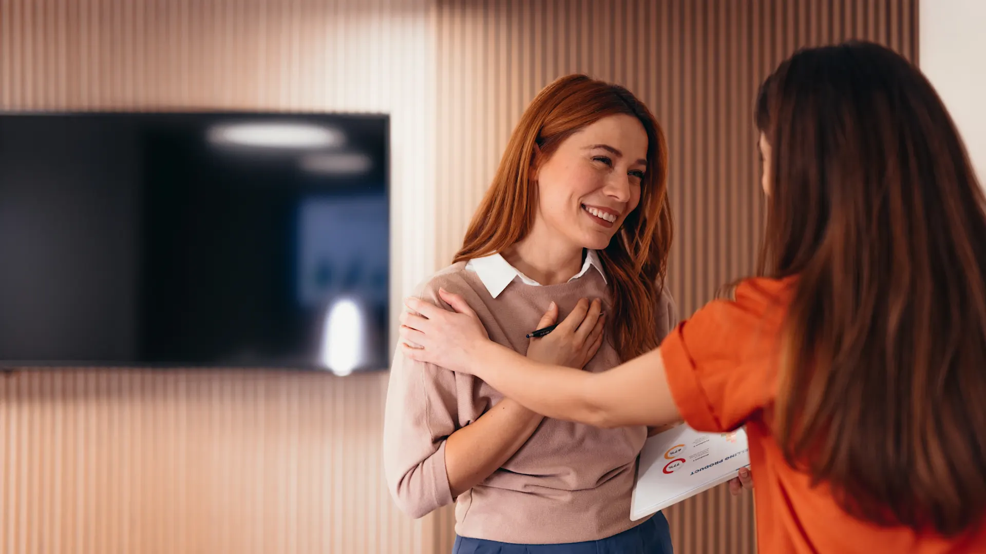 Mentale Gesundheit am Arbeitsplatz Zwei Frauen begrüßen sich herzlich in einem modern eingerichteten Raum, wobei eine rothaarige Frau in beigem Oberteil lächelnd ihre Gesprächspartnerin in orangefarbener Kleidung die Hand auf die Schulter legt.