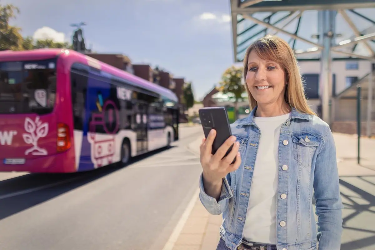 Frau steht an einer Bushaltestelle und schaut in ihr Smartphone. Ein NEW Bus ist im Hintergrund zu sehen.