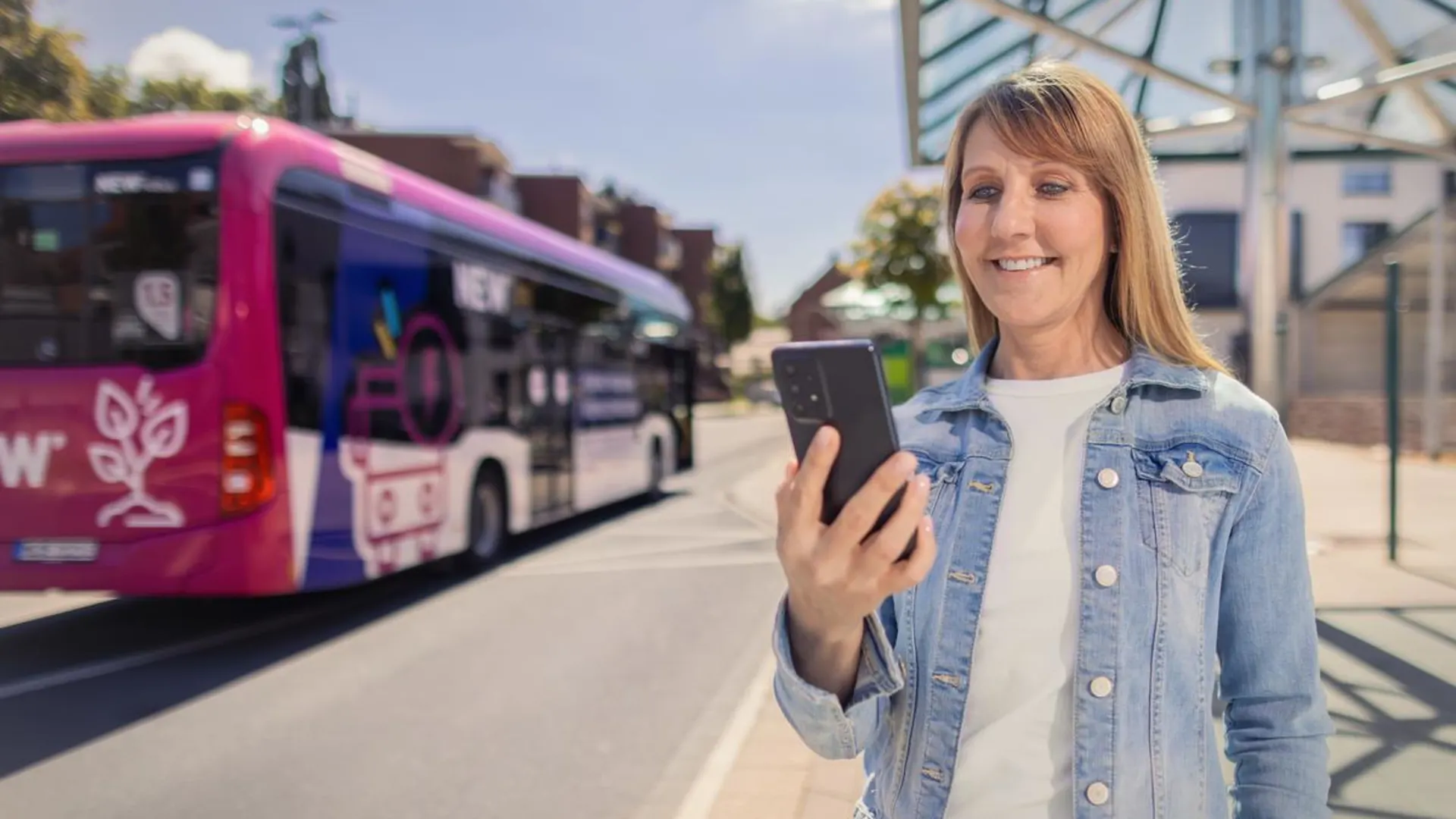 Tickets und Tarife Frau steht an einer Bushaltestelle und schaut in ihr Smartphone. Ein NEW Bus ist im Hintergrund zu sehen.