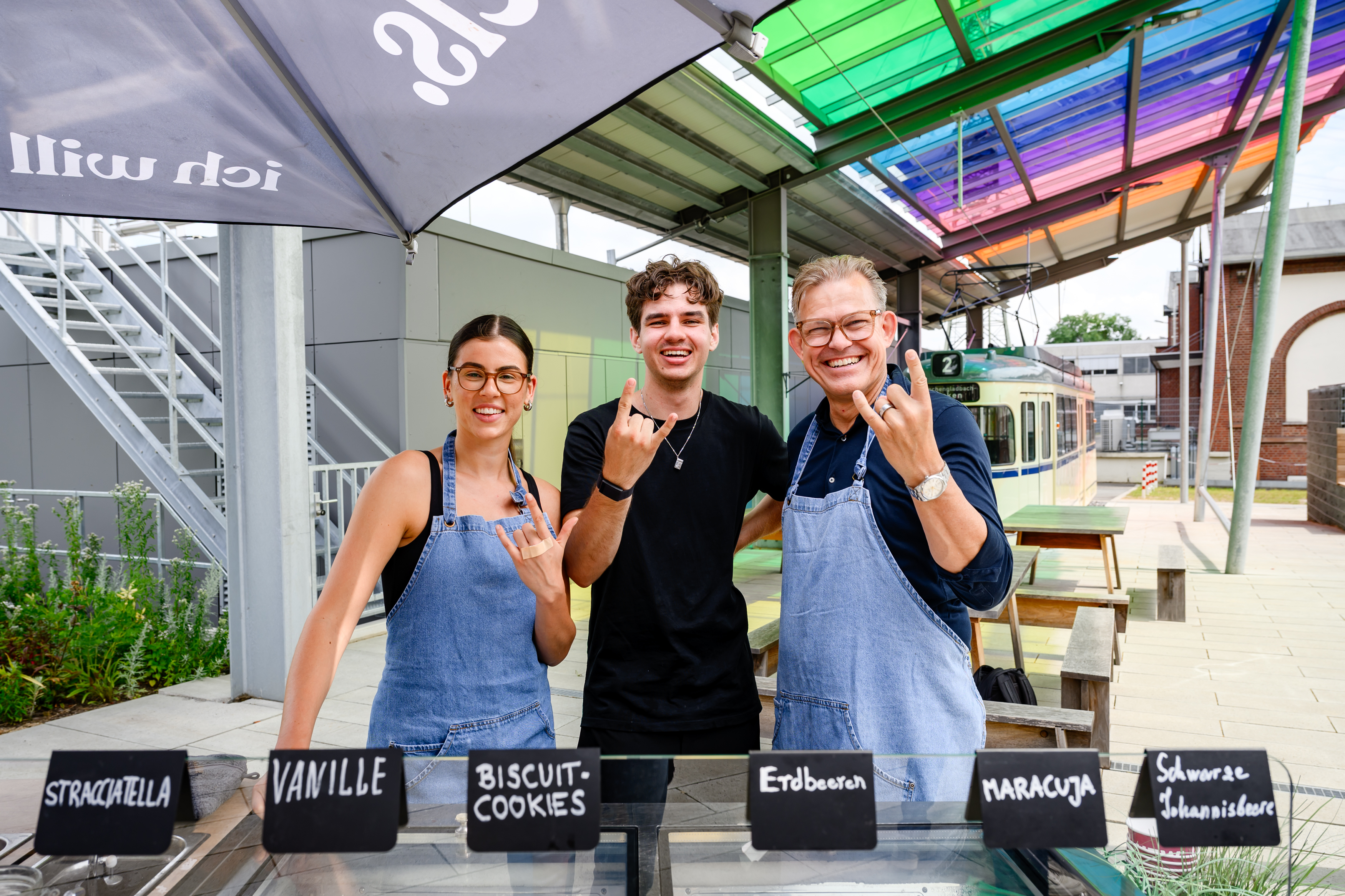 Drei Menschen stehen lächelnd hinter einem Eisverkaufsstand und zeigen ein Rock'n'Roll-Zeichen in die Kamera. Die beiden Personen in blauen Jeanslatzhosen flankieren einen Mann in schwarzem Shirt. Vor ihnen sind kleine schwarze Tafeln mit Eissorten wie Vanille, Erdbeeren und Maracuja aufgestellt, während im Hintergrund ein modernes Gebäude mit buntem, regenbogenfarbenem Glasdach und eine historische Straßenbahn zu sehen sind.