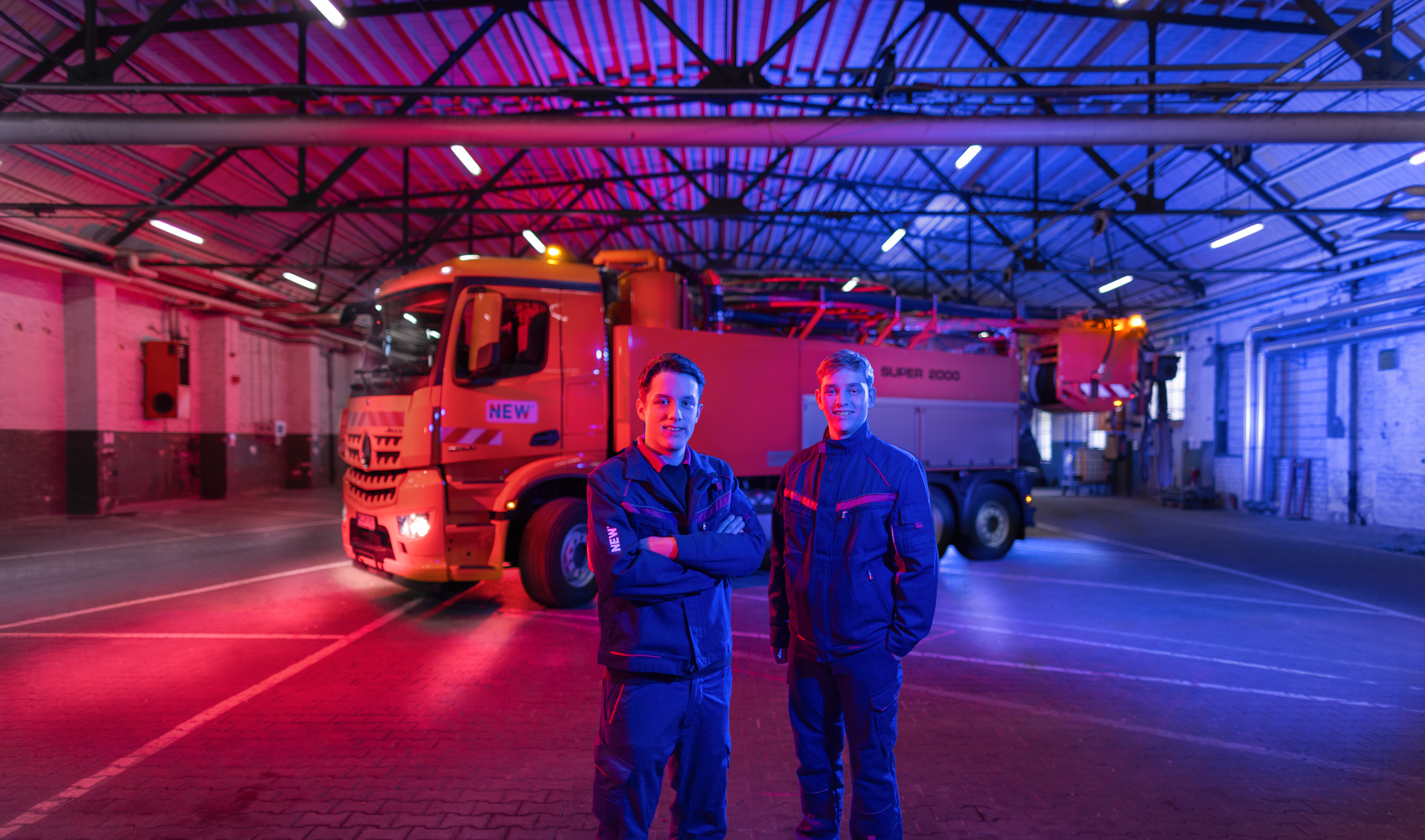 Industriehalle mit rot-blauer Beleuchtung, in der ein großer Lastwagen steht. Im Vordergrund sind zwei Personen in Arbeitskleidung zu sehen. 