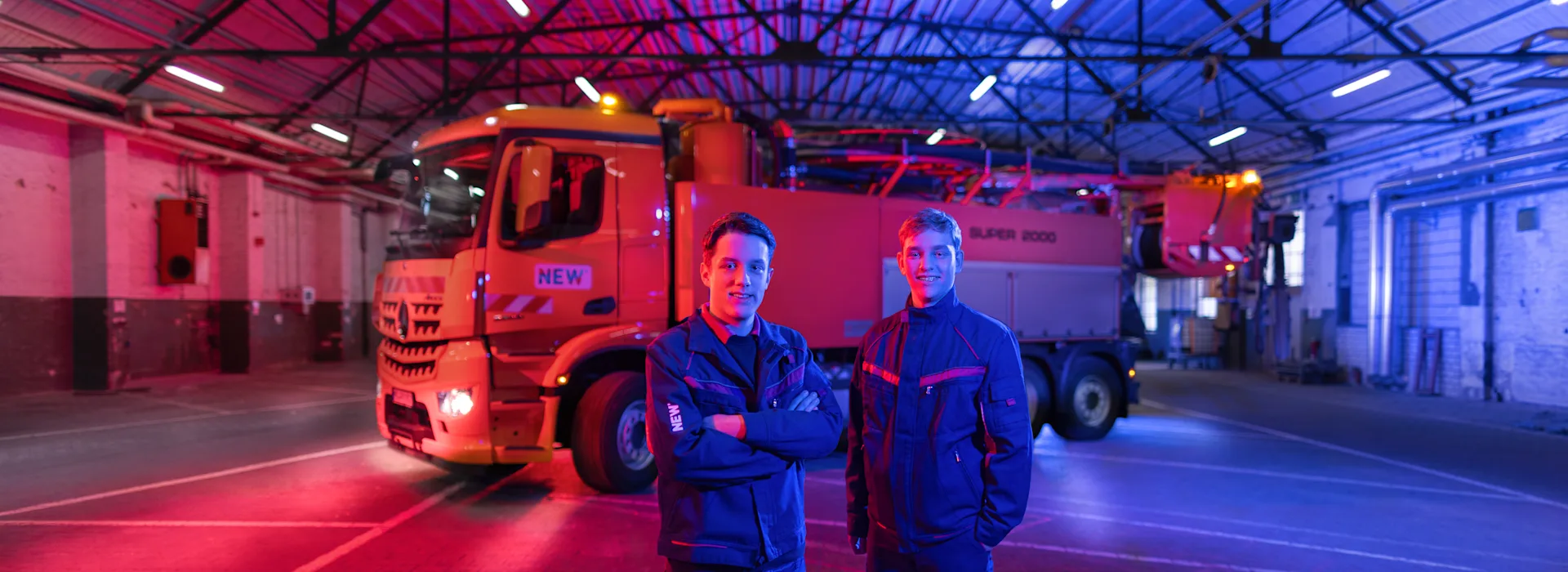 NEW Kollegen vor dem Super 2000 Industriehalle mit rot-blauer Beleuchtung, in der ein großer Lastwagen steht. Im Vordergrund sind zwei Personen in Arbeitskleidung zu sehen.