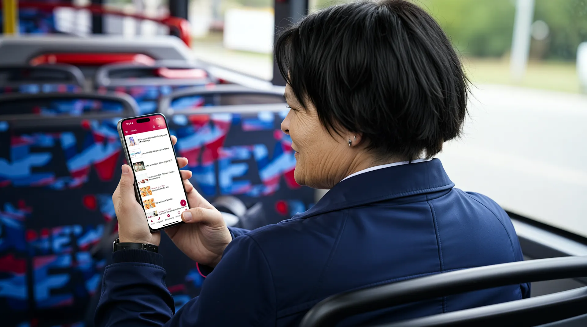 Eine Frau mit schwarzem Bob-Haarschnitt in dunkelblauer Jacke betrachtet eine Fahrplan-App auf ihrem Smartphone an einem Bus. Im Hintergrund sind Sitzrehen mit blau-rotem Muster zu sehen. Die Aufnahme zeigt die Nutzung digitaler Mobilitätsdienste im öffentlichen Nahverkehr.