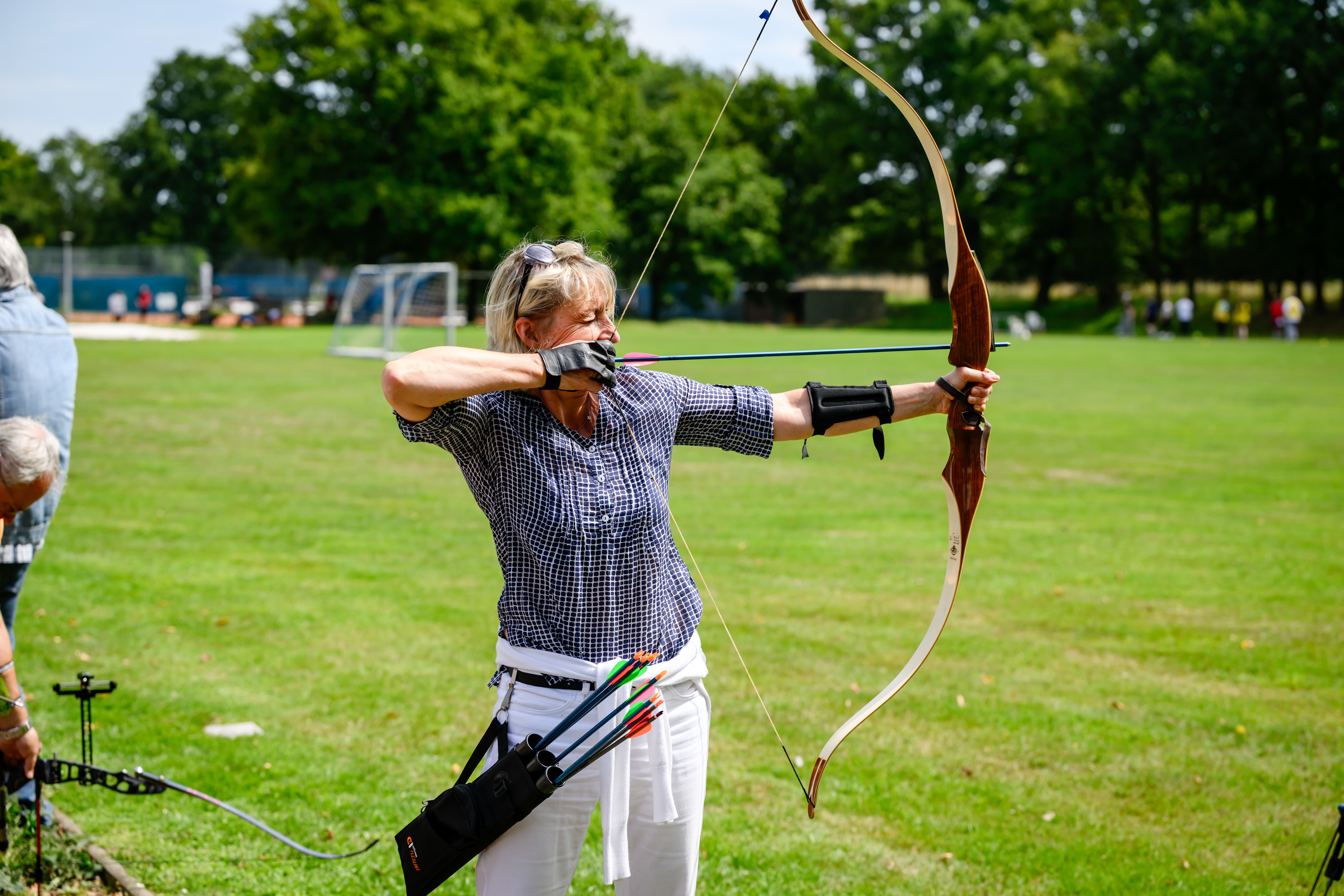 Eine Frau mit blonden Haaren in kariertem Hemd und weißer Hose zielt konzentriert mit einem Recurvebogen auf einem Sportplatz. Sie trägt einen schwarzen Armschutz und einen Köcher mit bunten Pfeilen am Gürtel. Im Hintergrund sind saftig grüne Bäume, Sportanlagen und weitere Personen zu sehen.