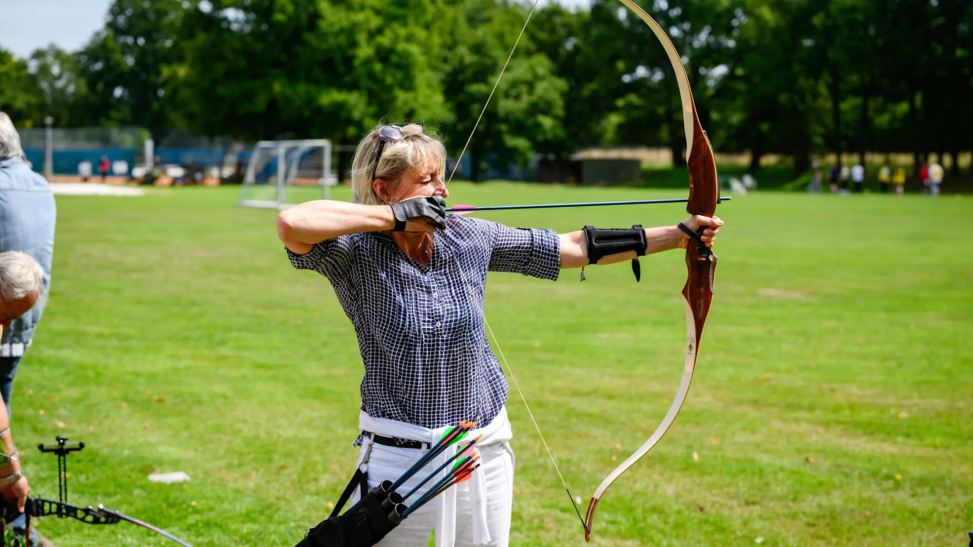 NEW Azubi Sport Tag Eine Frau mit blonden Haaren in kariertem Hemd und weißer Hose zielt konzentriert mit einem Recurvebogen auf einem Sportplatz. Sie trägt einen schwarzen Armschutz und einen Köcher mit bunten Pfeilen am Gürtel. Im Hintergrund sind saftig grüne Bäume, Sportanlagen und weitere Personen zu sehen.
