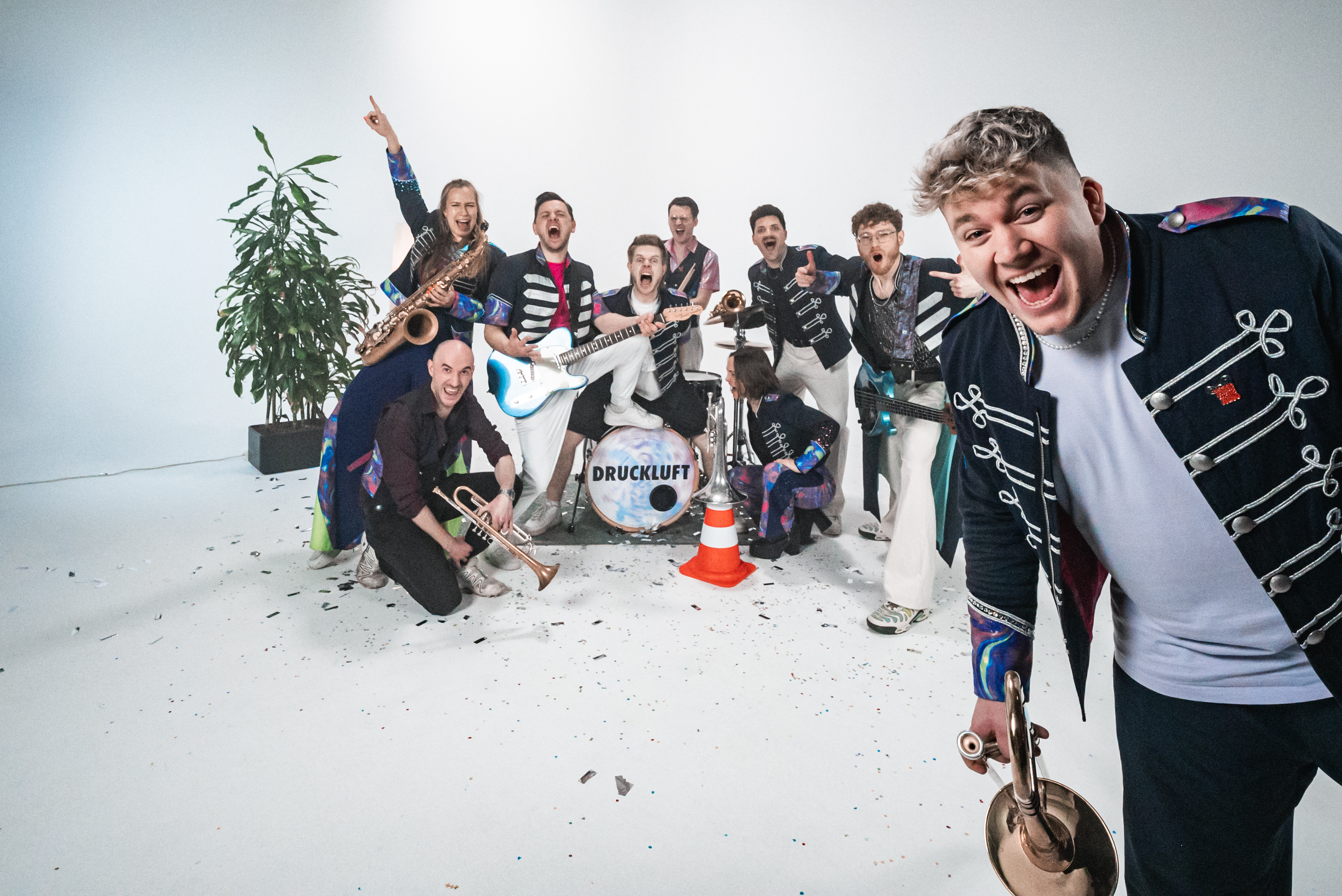 Die Band Druckluft präsentiert sich als energiegeladene Performance-Gruppe in einem hellen Studio-Setting. Mehrere Bandmitglieder stehen und knien rund um ein zentrales Schlagzeug mit Bandlogo, während unterschiedliche Blas- und Instrumente sichtbar sind. Im Vordergrund ist eine Person mit einem Blasinstrument nah zur Kamera positioniert. Die Outfits kombinieren dunkle Bühnenkleidung mit auffälligen, farbigen Details im Stil moderner Marchingbands. Pflanzen- und Dekoelemente ergänzen die Szene. Das Bild vermittelt Dynamik, Zusammenhalt und Live‑Musik mit hohem Entertainmentfaktor.