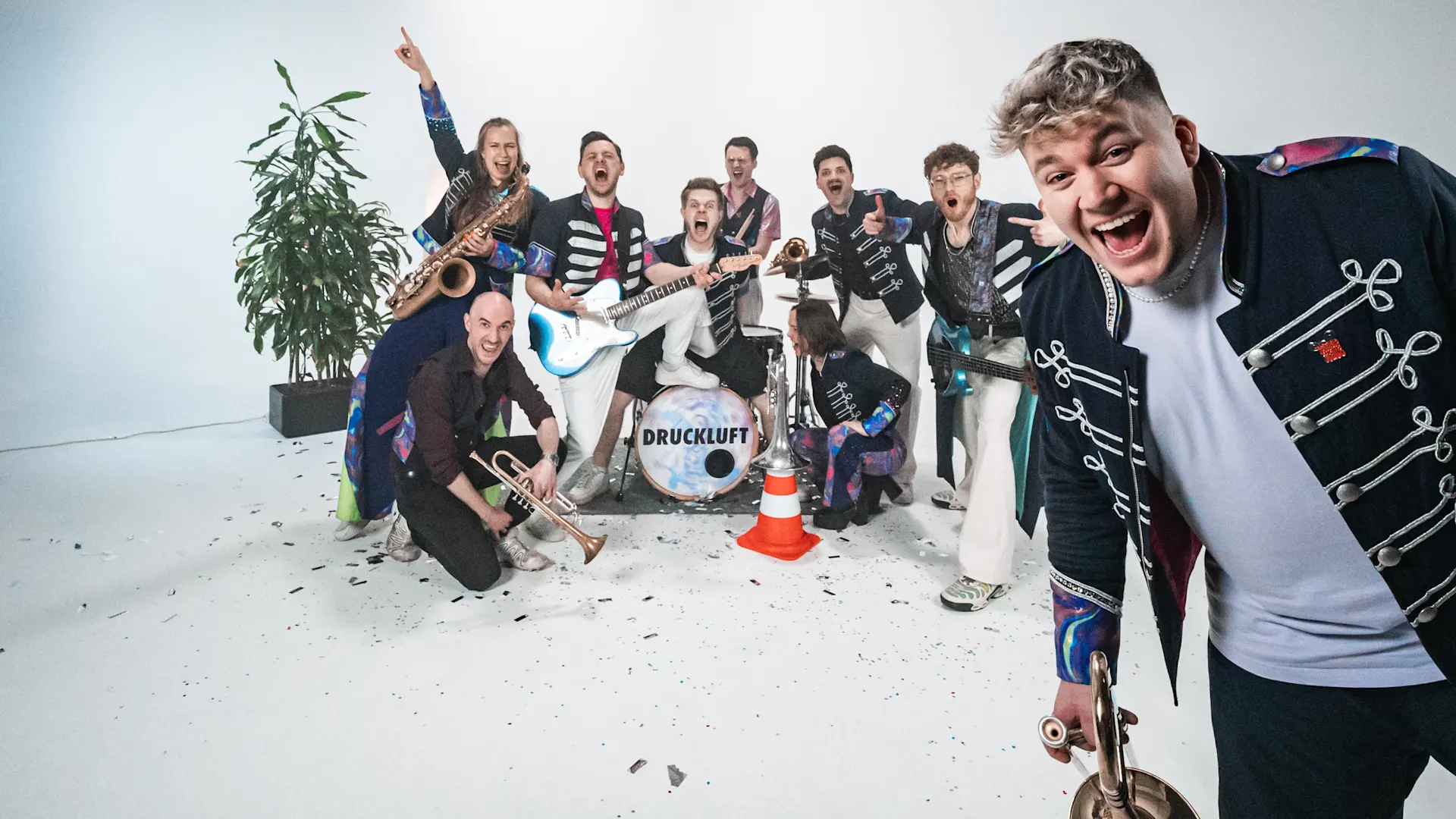 DRUCKLUFT (credits wewerekids) Die Band Druckluft präsentiert sich als energiegeladene Performance-Gruppe in einem hellen Studio-Setting. Mehrere Bandmitglieder stehen und knien rund um ein zentrales Schlagzeug mit Bandlogo, während unterschiedliche Blas- und Instrumente sichtbar sind. Im Vordergrund ist eine Person mit einem Blasinstrument nah zur Kamera positioniert. Die Outfits kombinieren dunkle Bühnenkleidung mit auffälligen, farbigen Details im Stil moderner Marchingbands. Pflanzen- und Dekoelemente ergänzen die Szene. Das Bild vermittelt Dynamik, Zusammenhalt und Live‑Musik mit hohem Entertainmentfaktor.