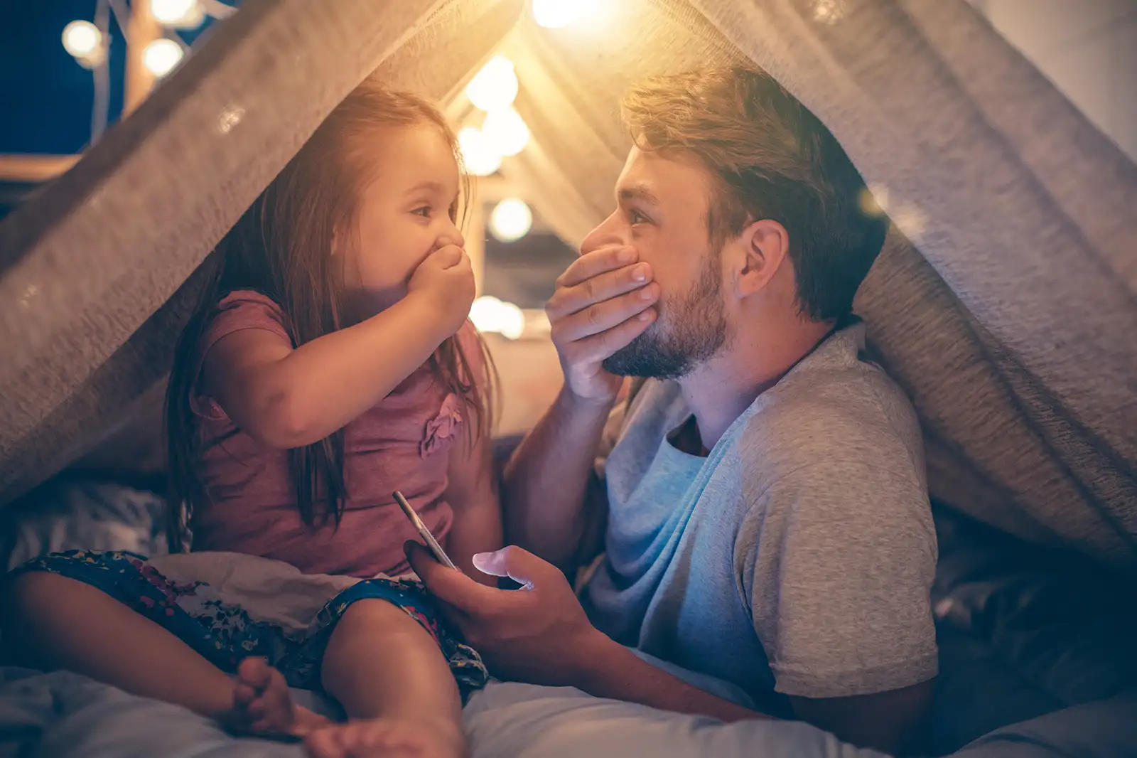 Vater und Tochter sitzen im Wohnzimmer unter einer Deckenhöhle und schauen gemeinsam aufs Smartphone und freuen sich über günstigen Strom.