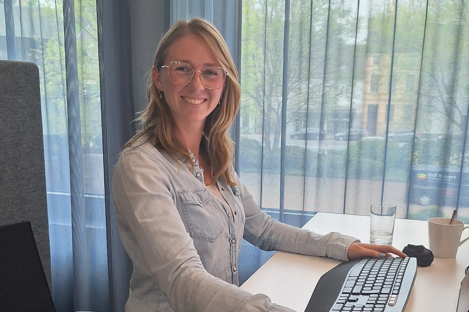 Person bei der Arbeit an einem Schreibtisch mit Computer vor einem Fenster mit durchscheinenden Vorhängen.