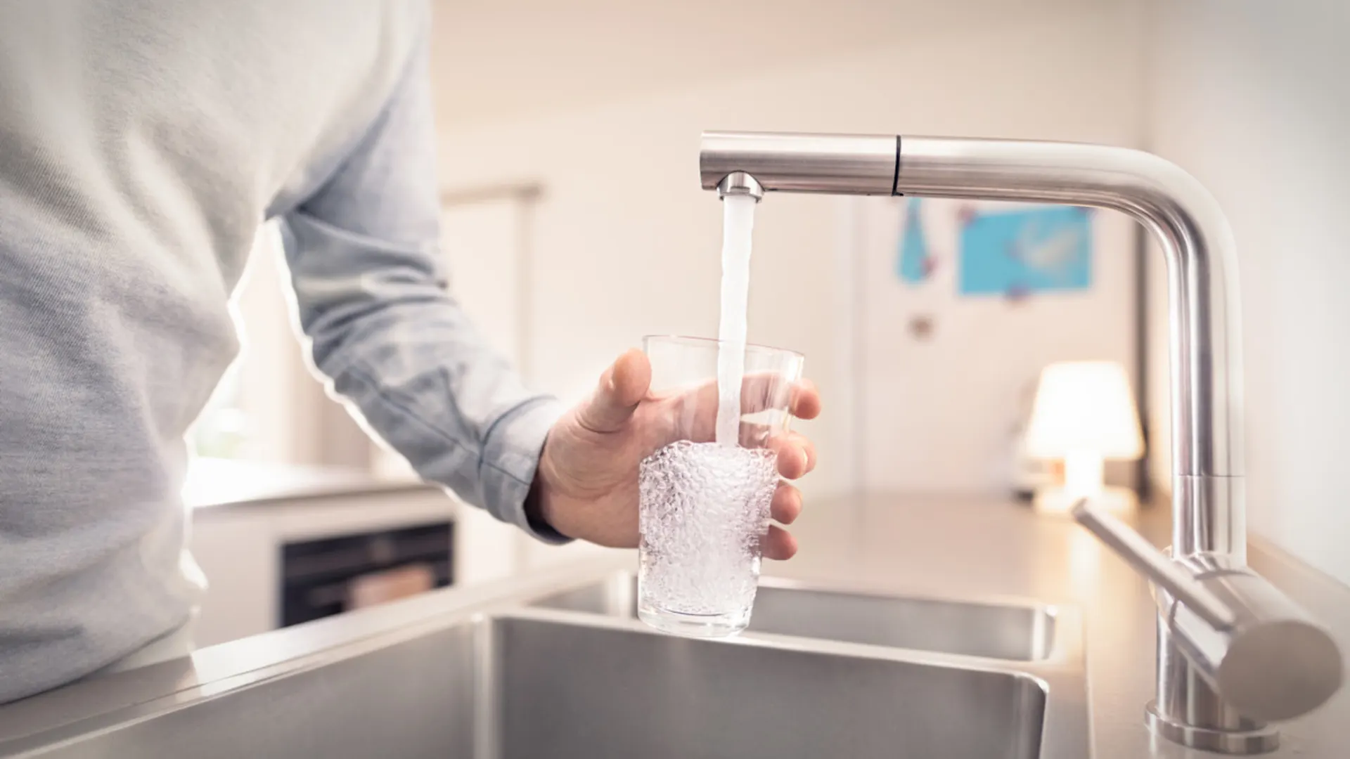 Ressourcen bewahren, Wasser schützen. Eine Person in hellem Pullover füllt ein Glas mit Wasser aus einem modernen Wasserhahn an einer Küchenspüle. Die Küche hat eine helle, moderne Ausstattung mit Edelstahlelementen und im Hintergrund sind Küchengeräte zu erkennen.
