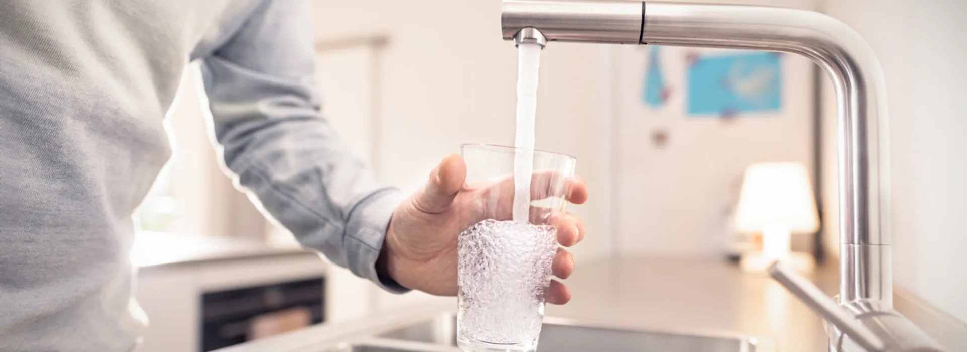 Ressourcen bewahren, Wasser schützen. Eine Person in hellem Pullover füllt ein Glas mit Wasser aus einem modernen Wasserhahn an einer Küchenspüle. Die Küche hat eine helle, moderne Ausstattung mit Edelstahlelementen und im Hintergrund sind Küchengeräte zu erkennen.