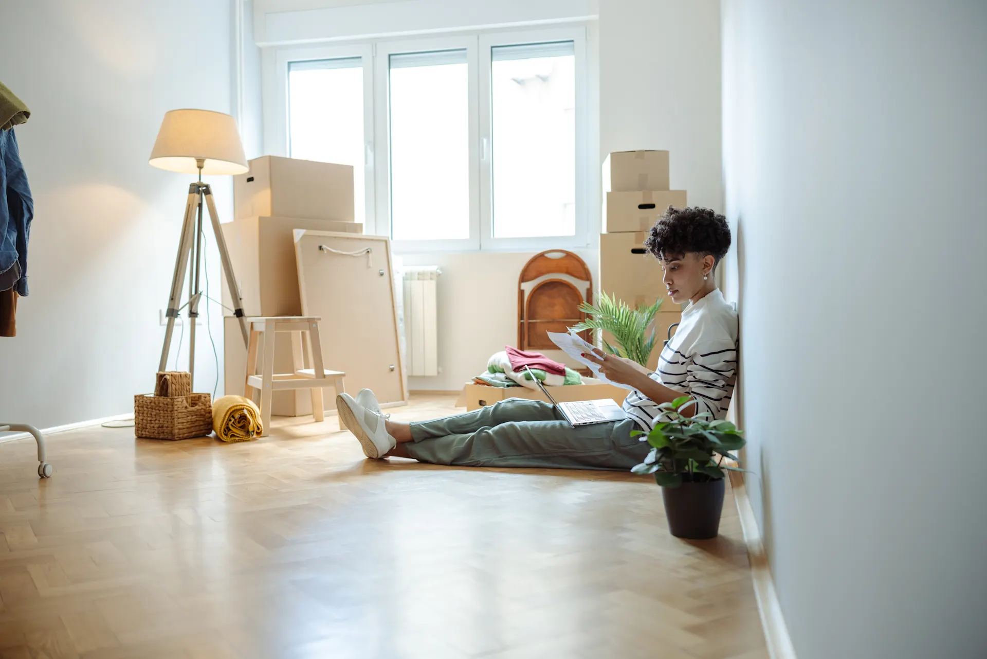 Erste eigene Wohnung - iStock/blackCAT Eine Person sitzt auf einem Holzboden in einem Zimmer mit Umzugskartons, liest Dokumente und hat einen Laptop auf dem Schoß.