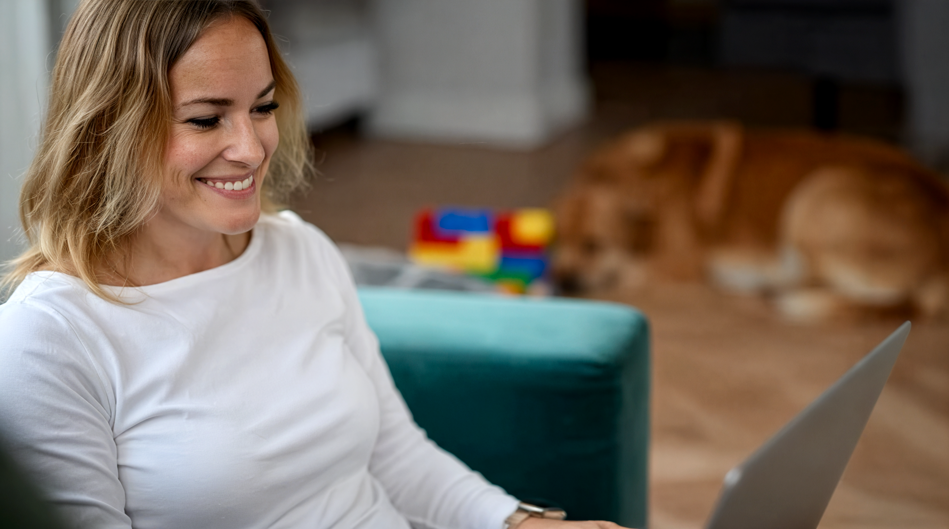Eine lächelnde Frau in weißem Langarmshirt arbeitet konzentriert an einem Laptop in einem hellen Wohnraum. Im Hintergrund sind buntes Spielzeug und ein Hund auf dem Holzboden zu sehen. Die Szene vermittelt eine entspannte Atmosphäre.