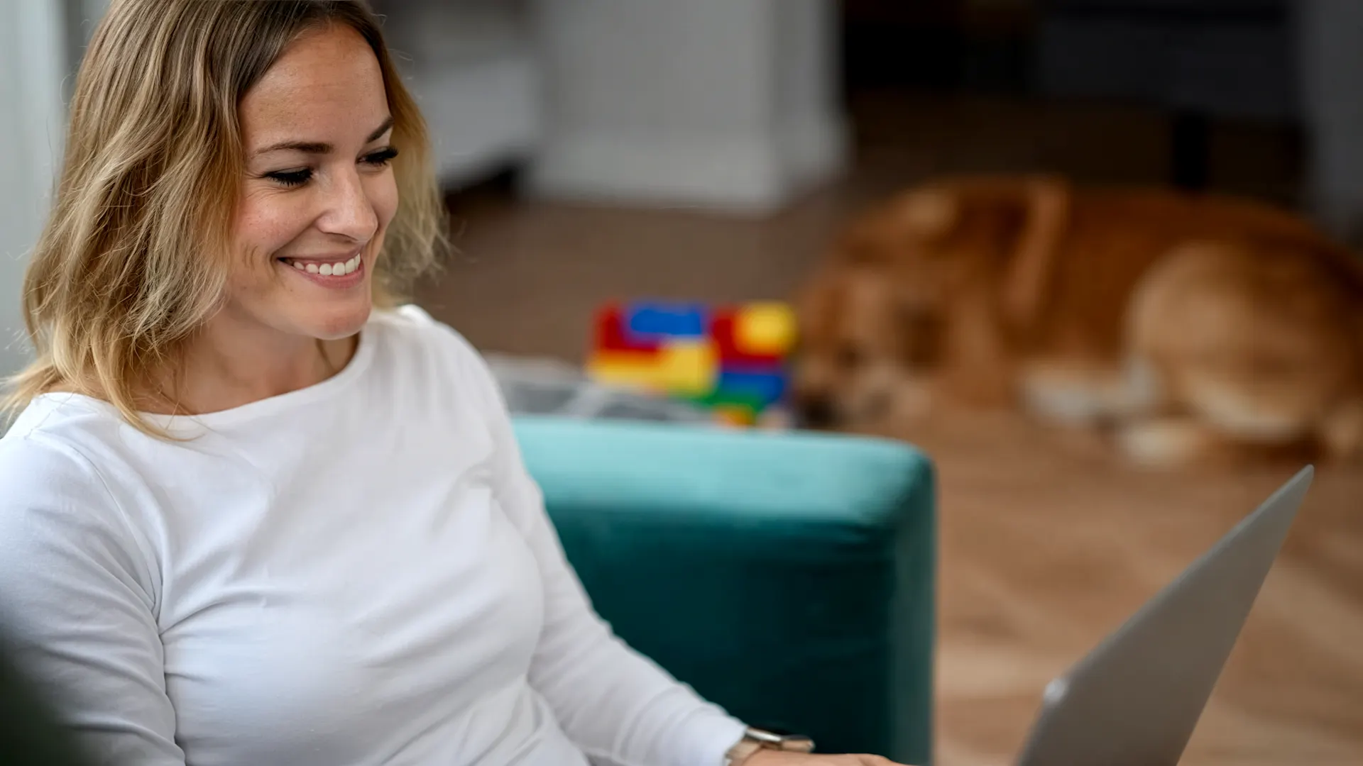 Bewerbungstipps Eine lächelnde Frau in weißem Langarmshirt arbeitet konzentriert an einem Laptop in einem hellen Wohnraum. Im Hintergrund sind buntes Spielzeug und ein Hund auf dem Holzboden zu sehen. Die Szene vermittelt eine entspannte Atmosphäre.