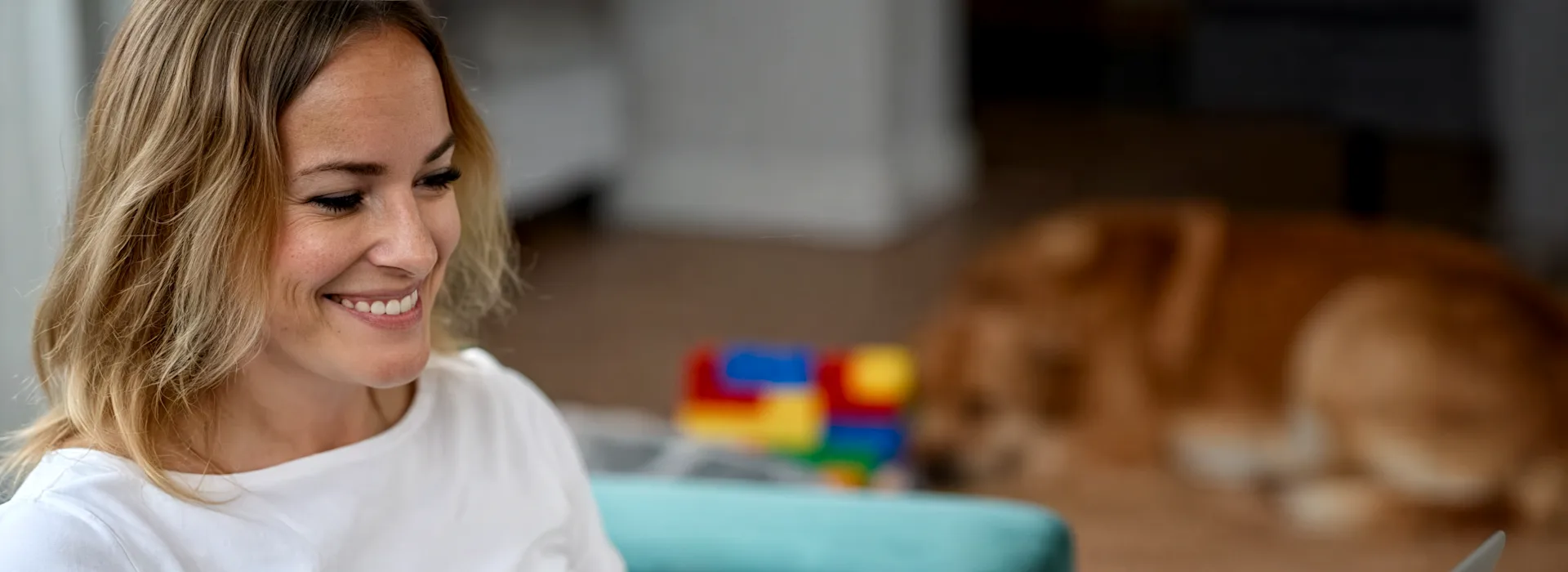Bewerbungstipps Eine lächelnde Frau in weißem Langarmshirt arbeitet konzentriert an einem Laptop in einem hellen Wohnraum. Im Hintergrund sind buntes Spielzeug und ein Hund auf dem Holzboden zu sehen. Die Szene vermittelt eine entspannte Atmosphäre.
