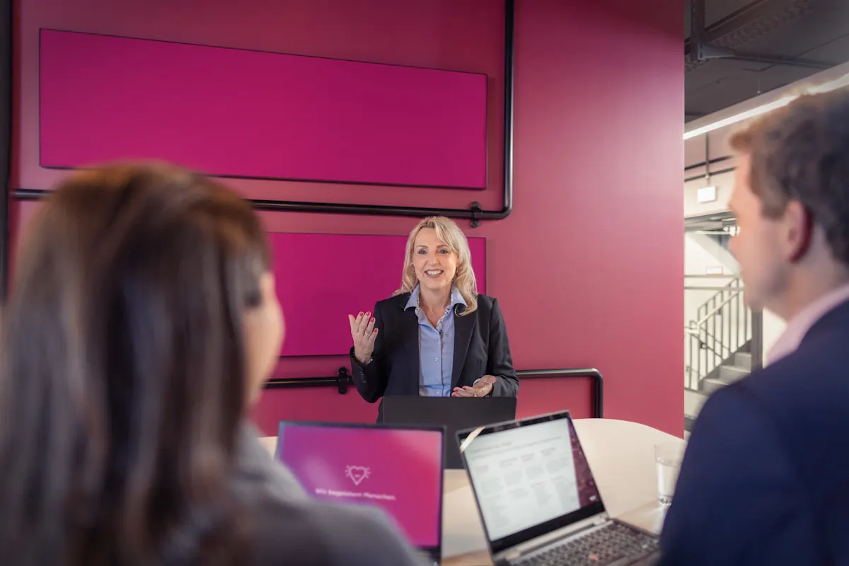 Ein Mann und zwei Frauen stehen zusammen an einem Tisch mit aufgeklappten Laptops und unterhalten sich.