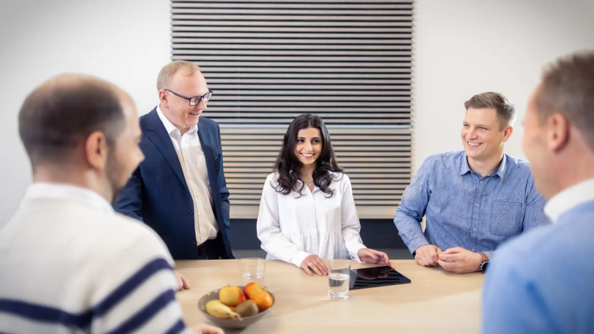 Rundum gut versorgt Eine Gruppe von fünf Kolleg*innen steht locker um einen Besprechungstisch und unterhält sich lächelnd in entspannter Atmosphäre. Auf dem Tisch liegen Obst, Wassergläser und ein Tablet.