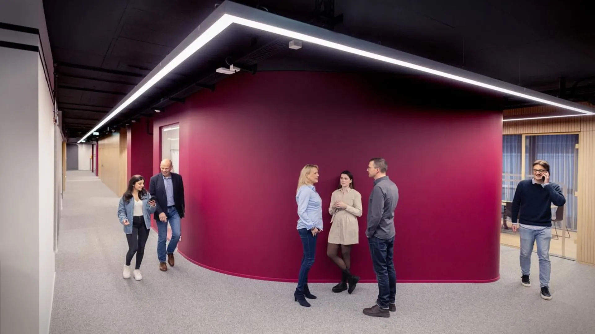 Arbeiten bei der NEW Mehrere Mitarbeitende der NEW unterhalten sich und gehen durch einen modernen Büroflur mit bordeauxroter Wand.