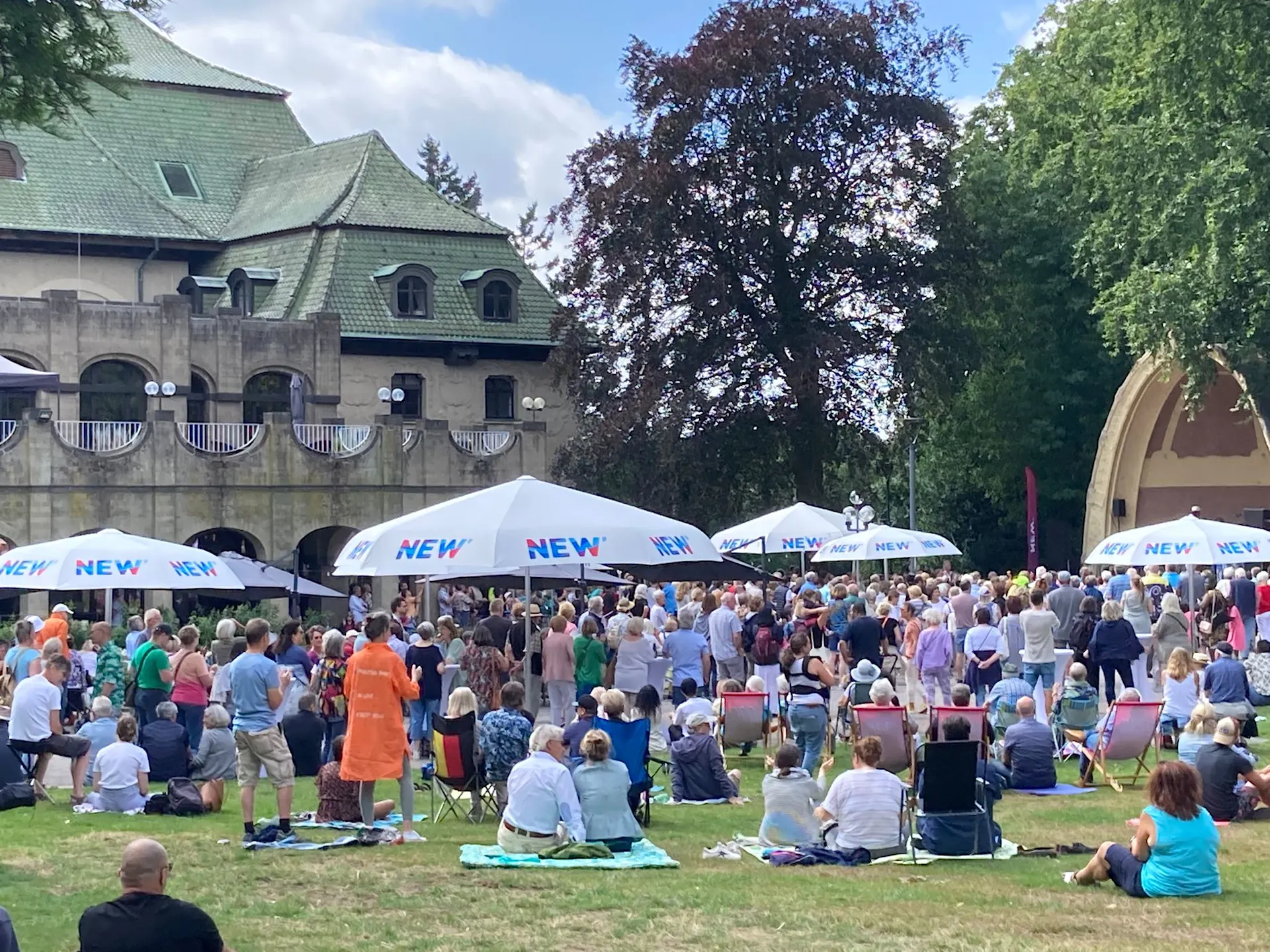 Eine große Menschenmenge versammelt sich bei einem Musikfestival im Freien vor einem Gebäude mit grünem Dach. Weiße Pavillons mit „NEW“ Beschriftung sind aufgebaut, und die Besucher:innen sitzen auf dem Rasen unter schattigen Bäumen.