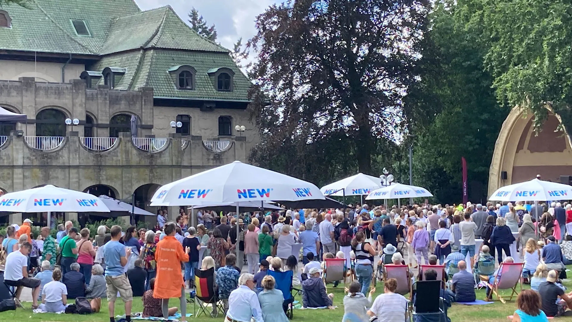 Musiksommer Eine große Menschenmenge versammelt sich bei einem Musikfestival im Freien vor einem Gebäude mit grünem Dach. Weiße Pavillons mit „NEW“ Beschriftung sind aufgebaut, und die Besucher:innen sitzen auf dem Rasen unter schattigen Bäumen.