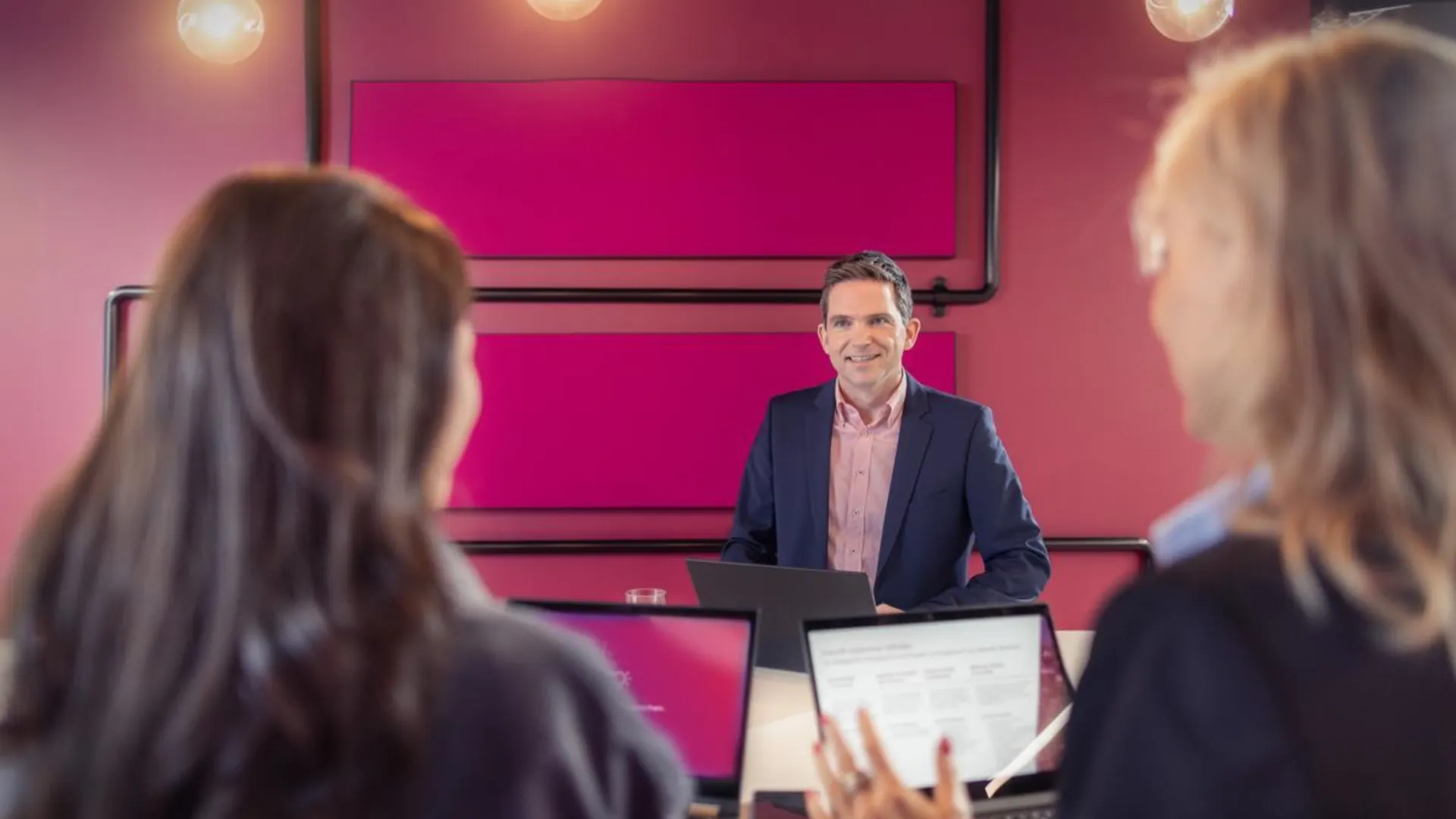Unternehmensleitbild bei der NEW Ein Mann im Anzug steht lächelnd an einem Besprechungstisch mit Laptop, zwei Kolleginnen mit geöffneten Laptops im Vordergrund hören ihm zu. Die Szene findet in einem modern gestalteten Raum mit pinker Wand statt.