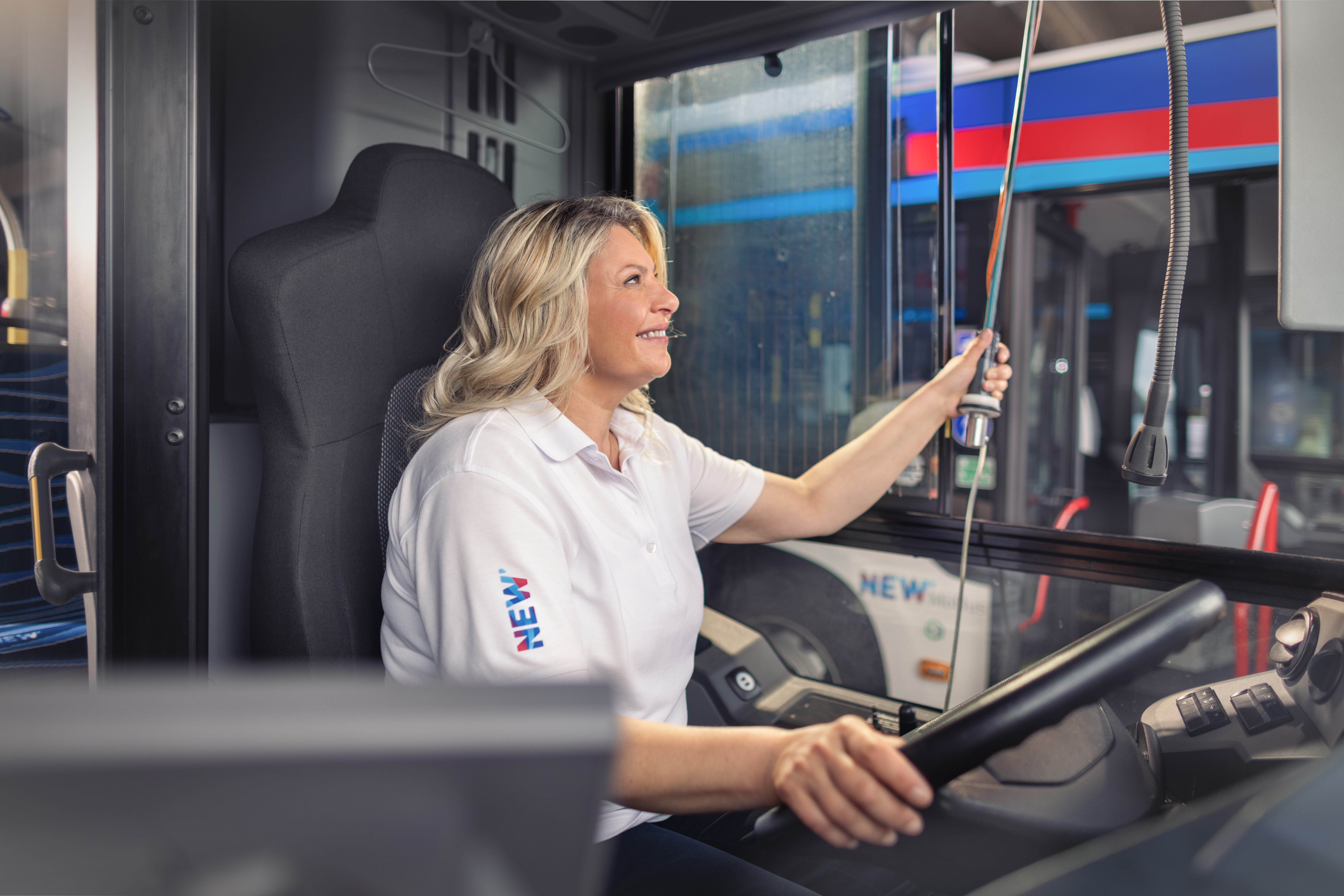 Eine Busfahrerin in weißer Uniform mit NEW-Logo entkoppelt ihr Fahrzeugs, während sie hinter dem Lenkrad sitzt. Im Hintergrund ist ein weiterer Bus in einer Fahrzeughalle zu sehen.