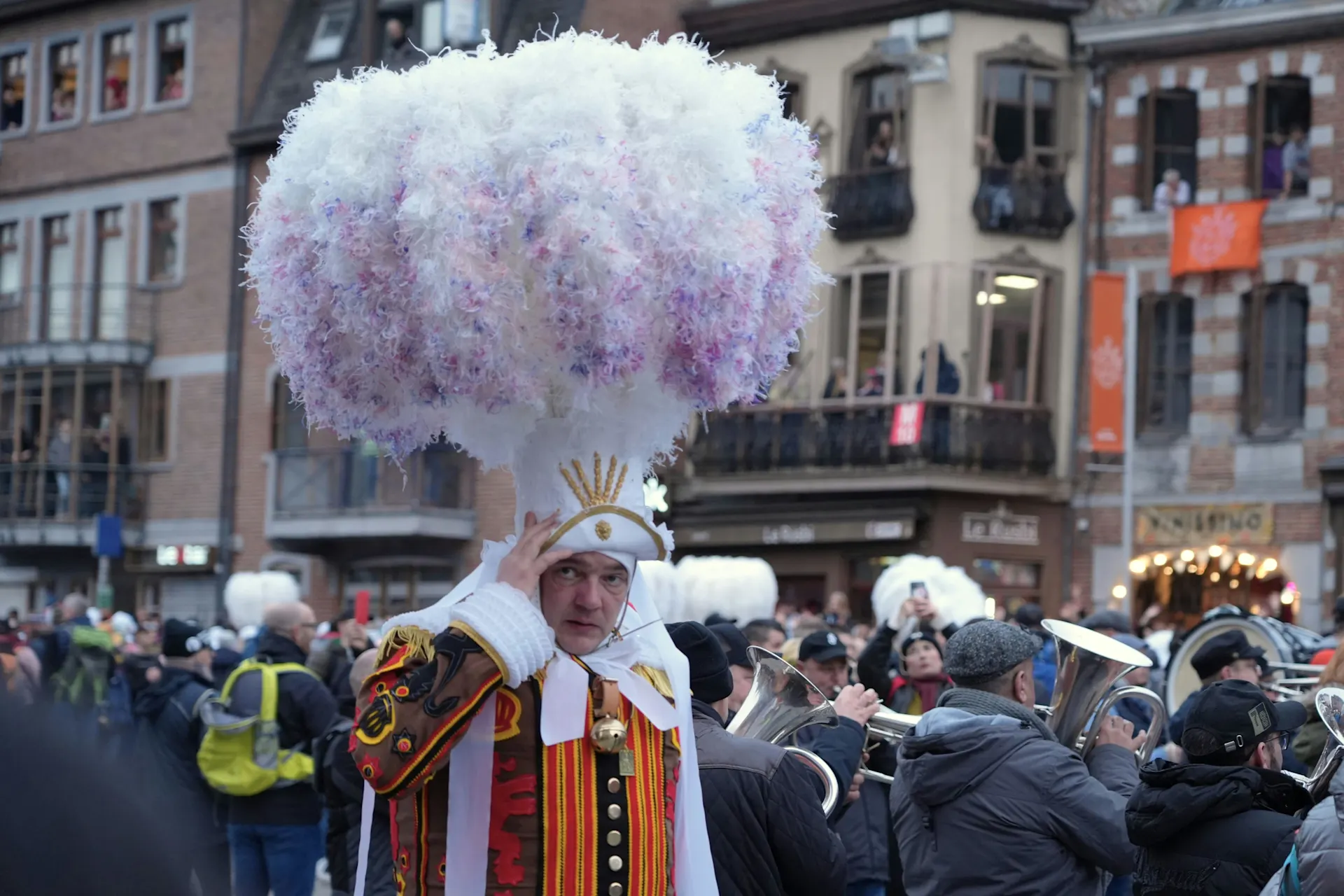 Ein Mann in rot-goldener Uniform mit weißen Akzenten und weißer Mütze mit goldener Krone. Besonders auffällig ist sein Kopfschmuck: eine große Perücke mit weiß-rosa-violetten Federn.