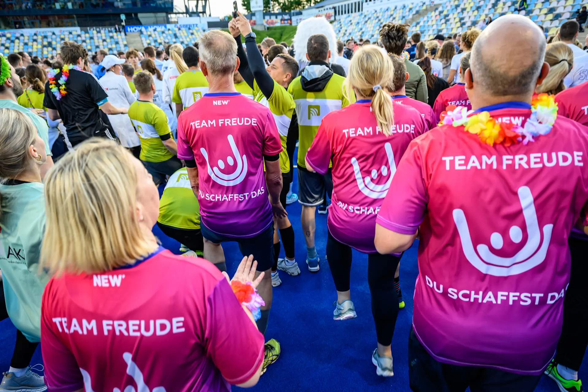 Läufer in pinken und lilafarbenen TEAM FREUDE-Shirts mit Smiley-Logo und der Aufschrift DU SCHAFFST DAS versammeln sich am Start eines Laufevents in einem Stadion. Im Hintergrund sind Zuschauerränge zu sehen.