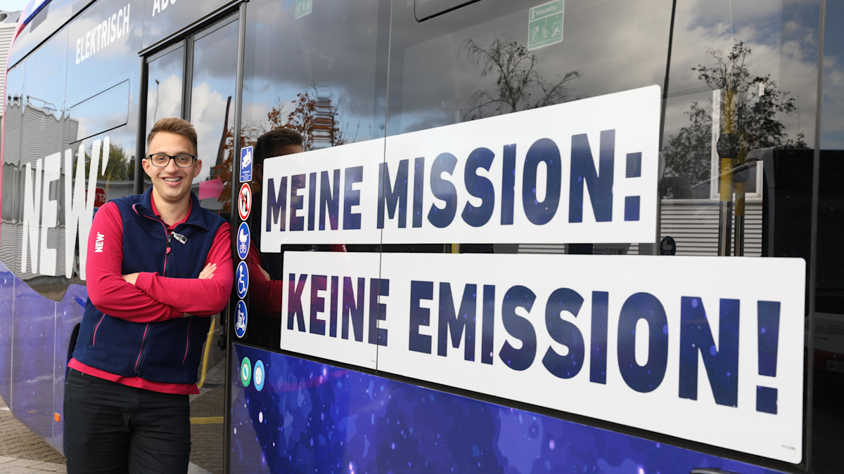 NEW Mitarbeiter steht angelehnt an einem neuen NEW E-Bus neben dem großen Aufkleber "Meine Mission: Keine Emission!"