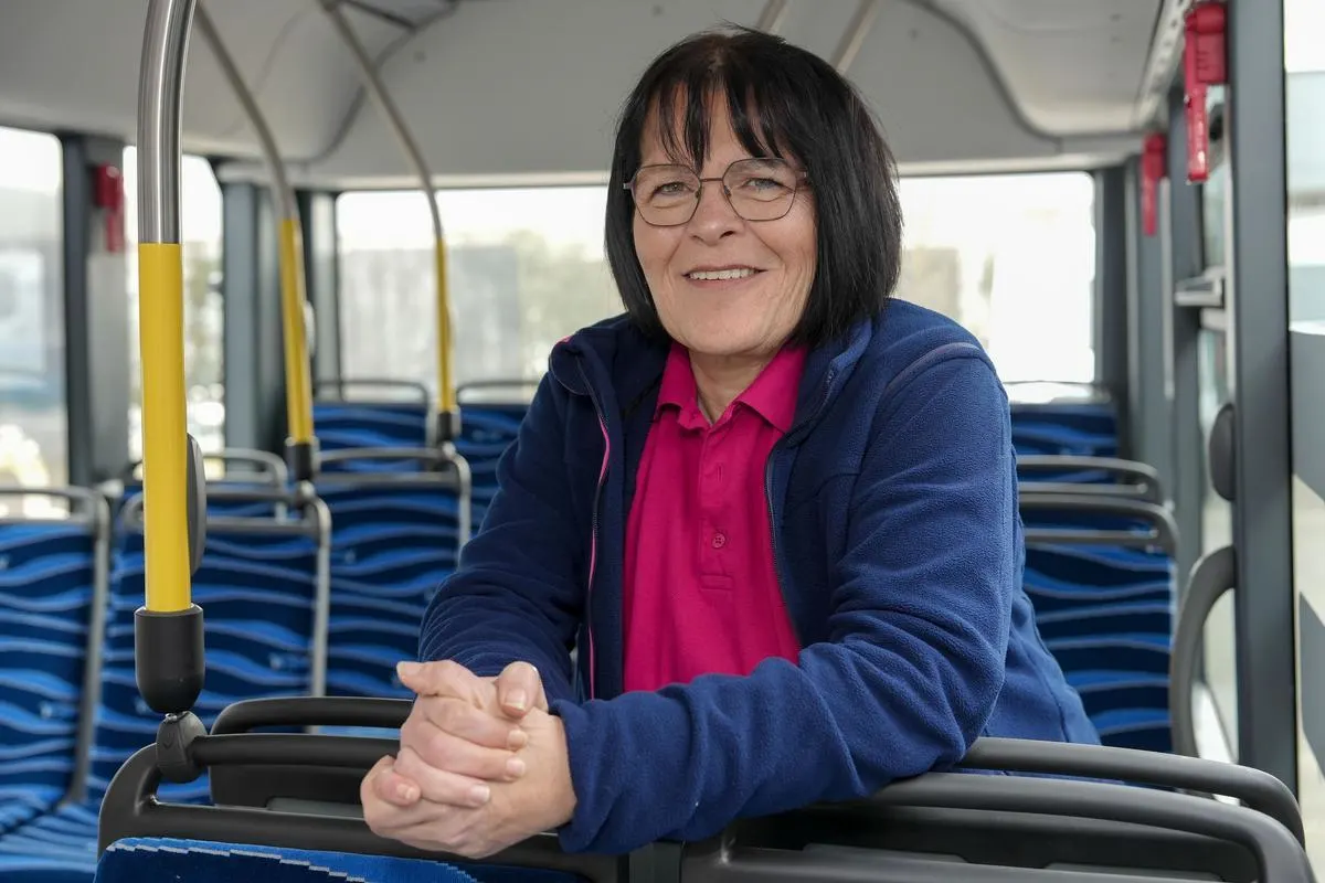 Eine lächelnde Busfahrerin der NEW sitzt in einem leeren Linienbus und blickt freundlich in die Kamera. Sie trägt eine dunkelblaue Jacke über einem pinken Poloshirt.