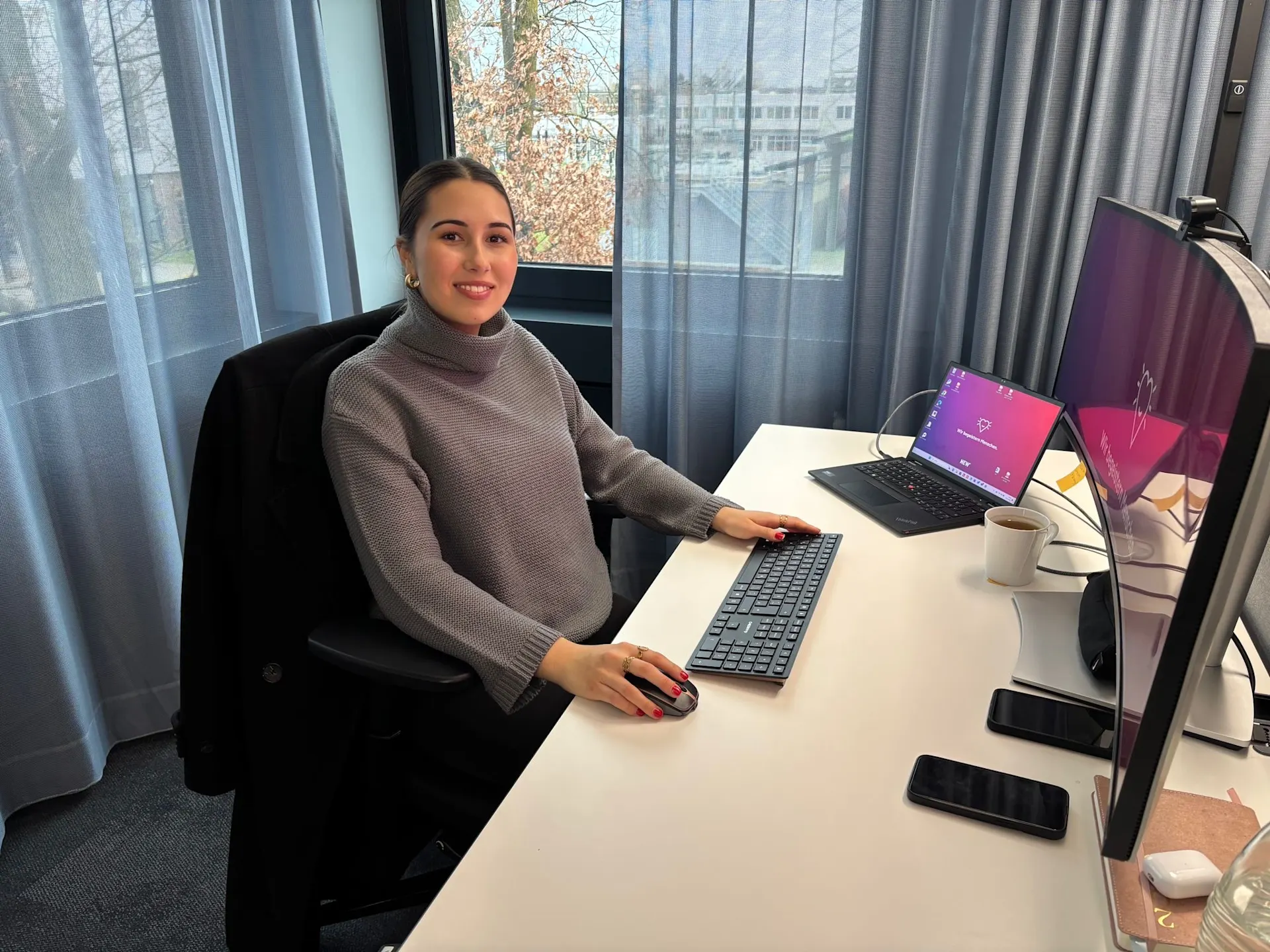 Junge Frau die an einem Schreibtisch sitzt und in die Kamera lächelt. Sie bedient eine Maus und hat vor sich ihren Laptop und einen großen Bildschirm stehen.