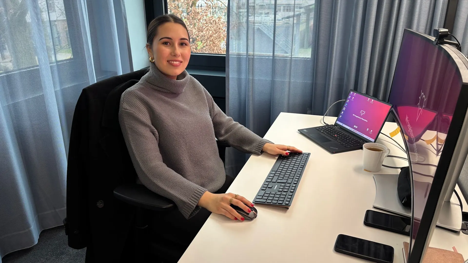 Duale Studentin bei der NEW - Victoria Khalil Eine junge Frau sitzt lächelnd an einem modernen Büroarbeitsplatz vor einem großen Monitor und einem Laptop. Sie arbeitet in einer hellen, freundlichen Umgebung mit Blick nach draußen. Auf dem Schreibtisch liegen Smartphone, Tasse und weitere Arbeitsmaterialien