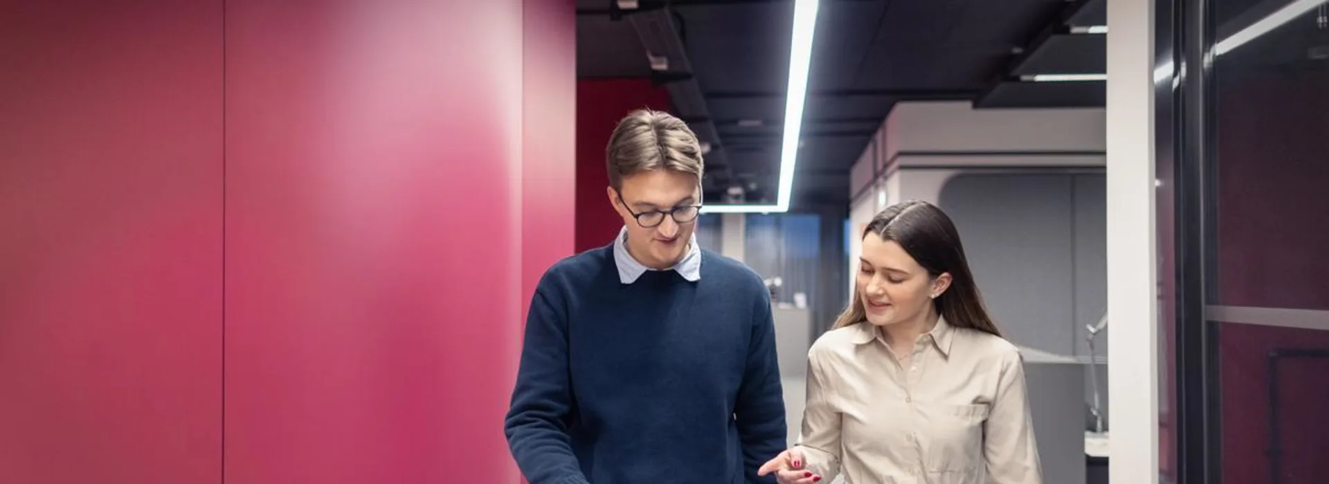 Studierende bei der NEW Zwei junge Erwachsene gehen gemeinsam durch einen modernen Büroflur und schauen dabei auf ein Tablet. Eine Person trägt das Tablet, die andere deutet mit dem Finger darauf.
