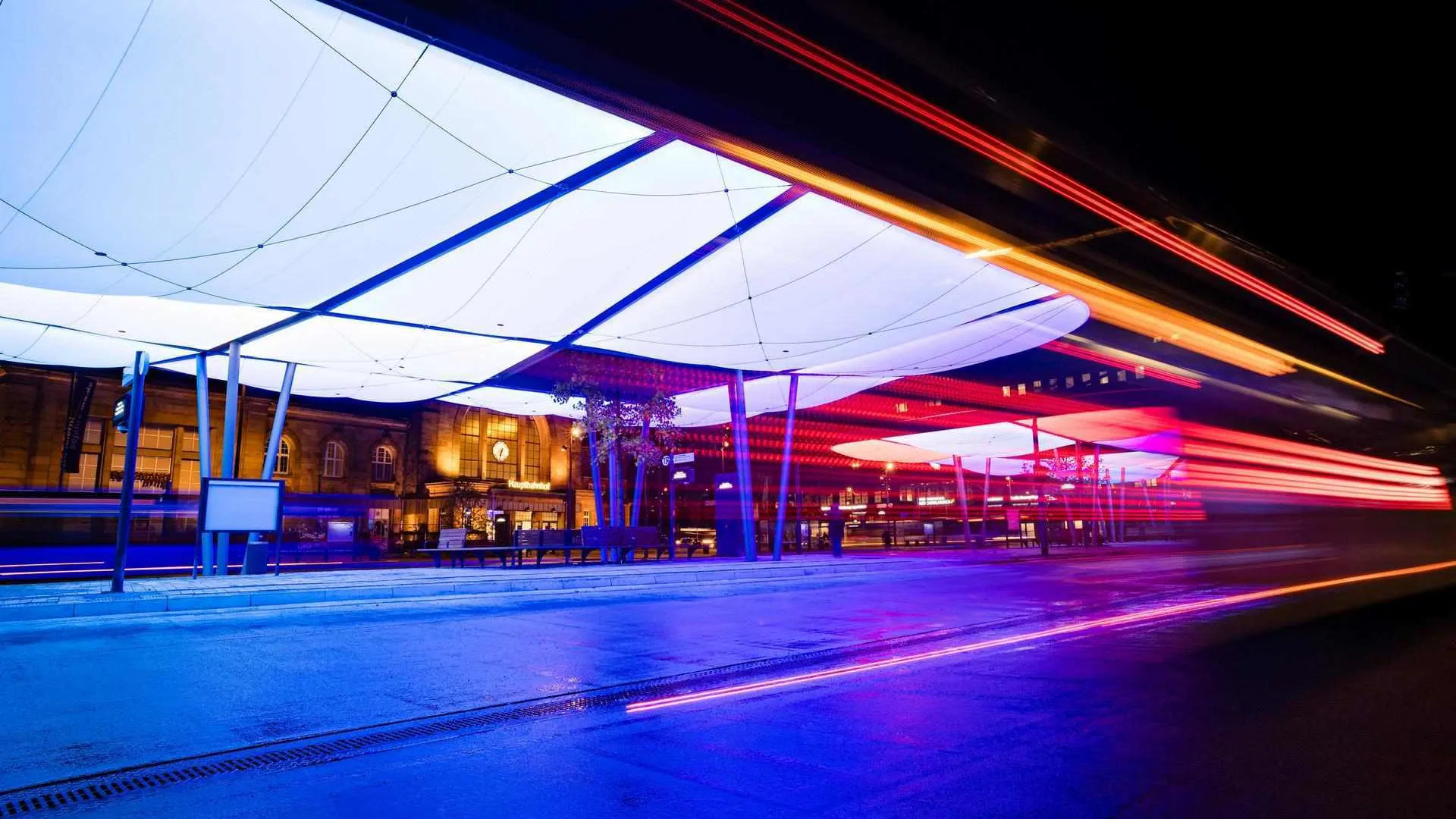 Busbahnhof-8 Das Bild zeigt den Zentralen Omnibusbahnhof (ZOB) in Mönchengladbach bei Nacht. Die moderne Dachkonstruktion ist farbig beleuchtet und prägt das Stadtbild. Im Hintergrund befindet sich der Hauptbahnhof. Die Szene wirkt dynamisch durch Lichtspuren vorbeifahrender Fahrzeuge.