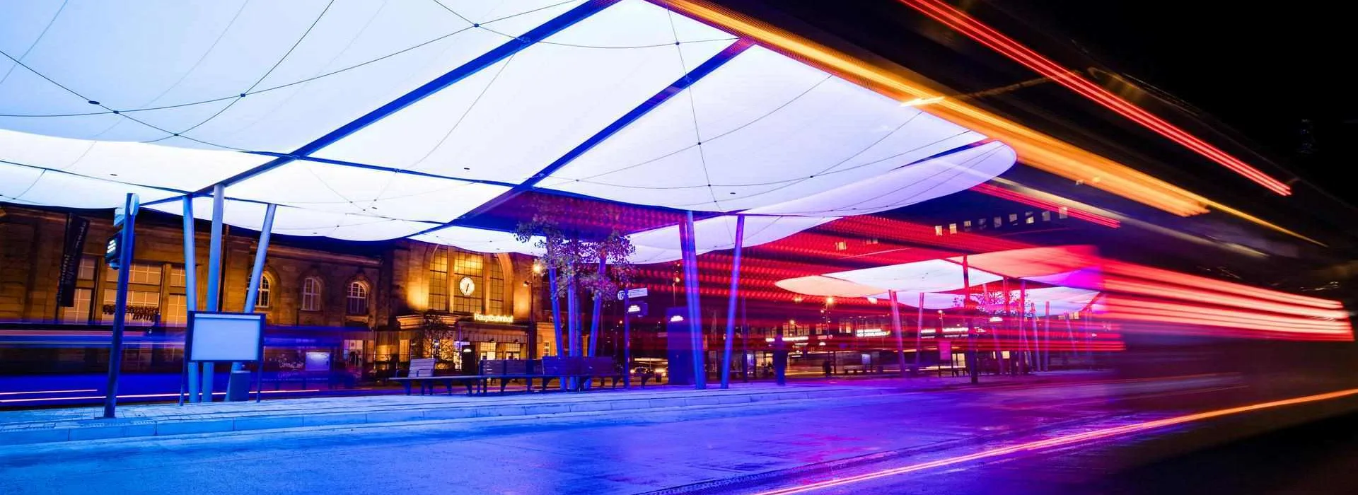 Busbahnhof-8 Das Bild zeigt den Zentralen Omnibusbahnhof (ZOB) in Mönchengladbach bei Nacht. Die moderne Dachkonstruktion ist farbig beleuchtet und prägt das Stadtbild. Im Hintergrund befindet sich der Hauptbahnhof. Die Szene wirkt dynamisch durch Lichtspuren vorbeifahrender Fahrzeuge.