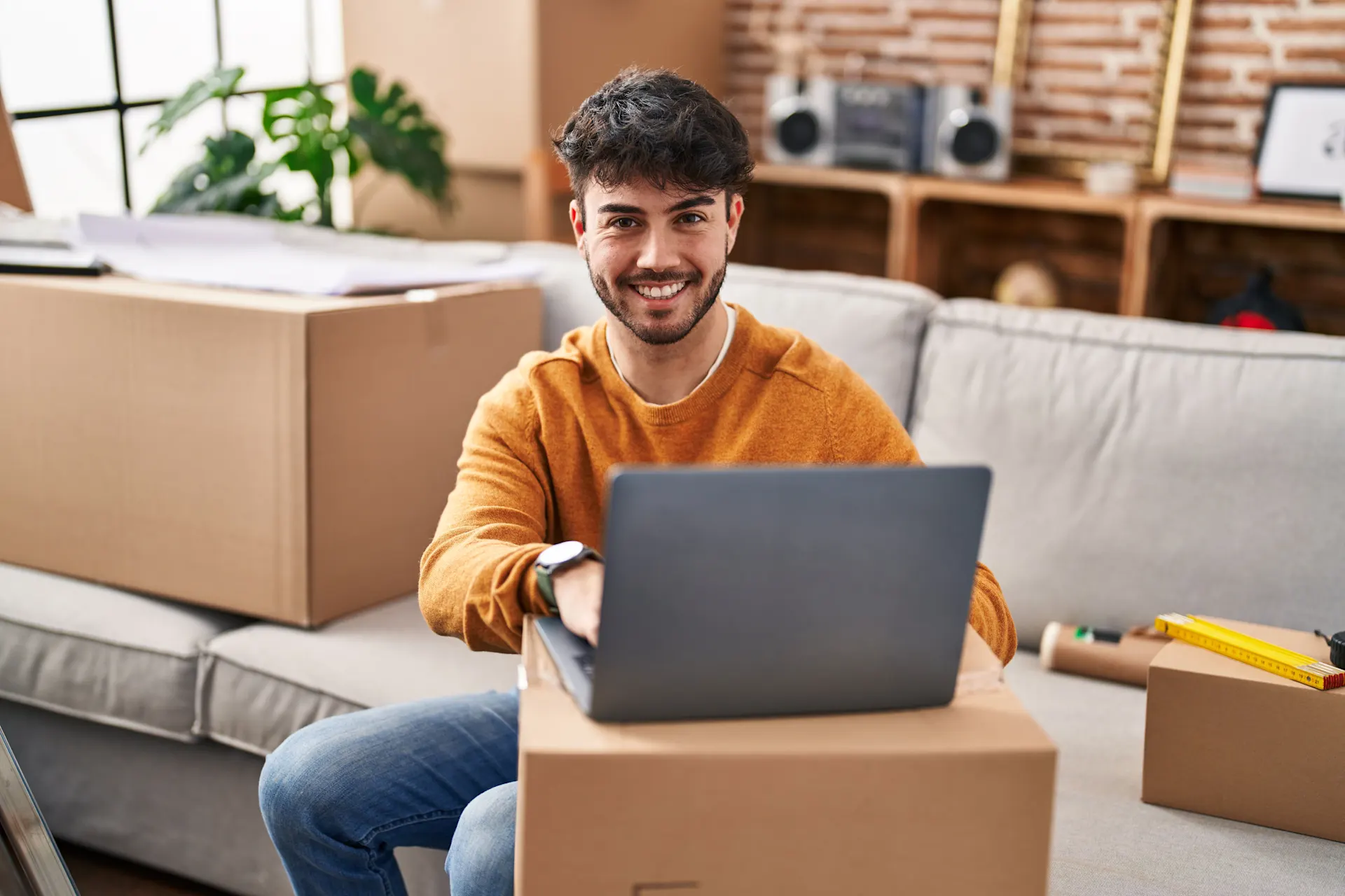 Guenstiger-Strom-und-Gas-fuer-Ihr-neues-Zuhause Ein junger Mann sitzt lächelnd zwischen Umzugskartons auf dem Sofa und hat einen Laptop vor sich stehen.