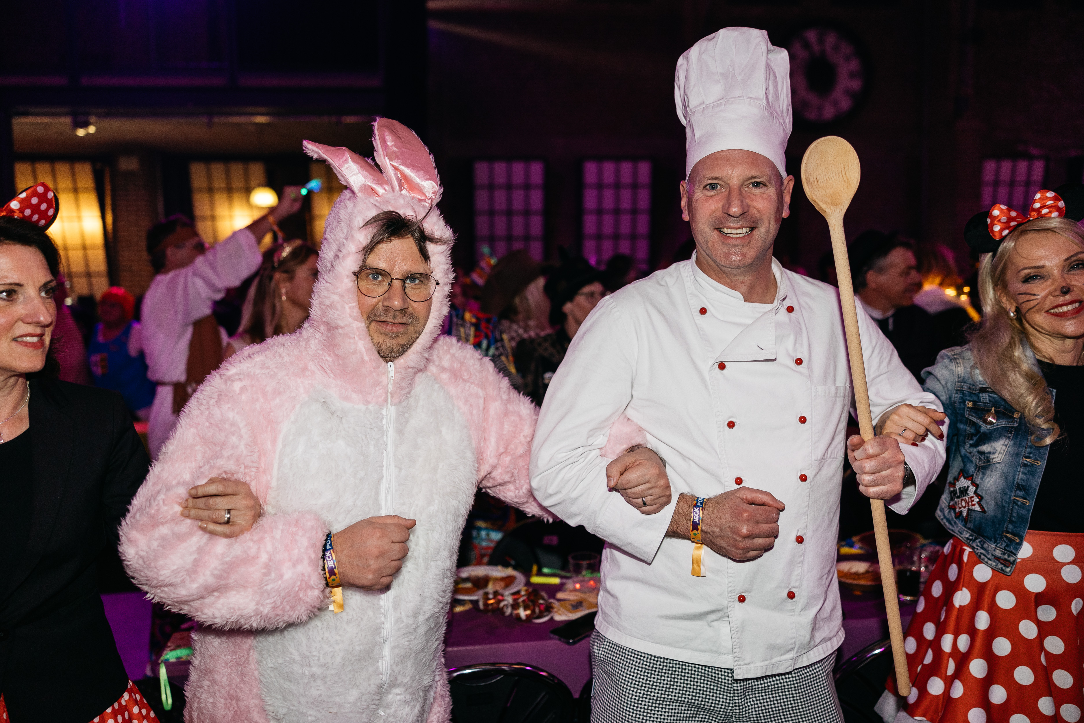 Mann in rosa Hasenkostüm steht Arm in Arm mit Mann in weißer Kochuniform und Kochmütze, der einen Holzkochlöffel hält. Beide lächeln in die Kamera, im Hintergrund ein festlich beleuchteter Raum mit weiteren Gästen. 