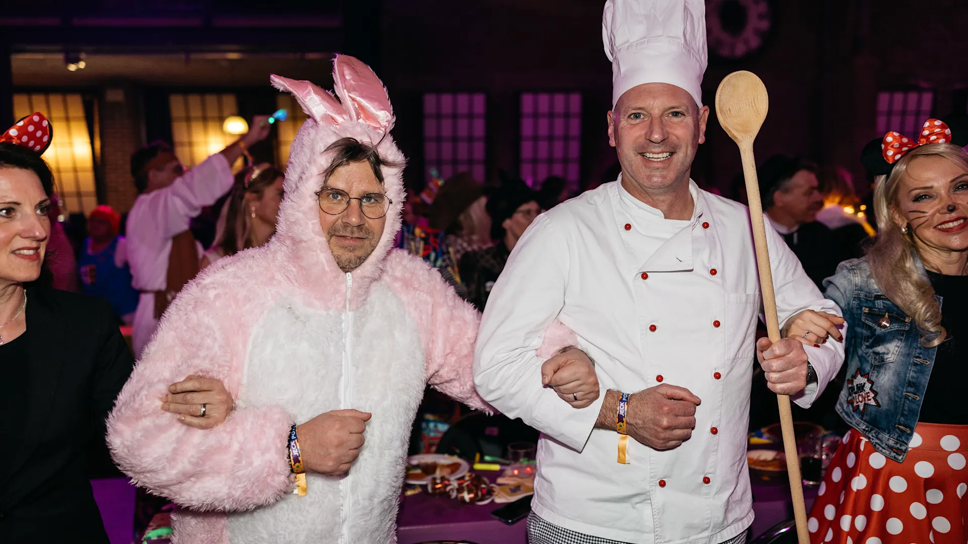Mann in rosa Hasenkostüm steht Arm in Arm mit Mann in weißer Kochuniform und Kochmütze, der einen Holzkochlöffel hält. Beide lächeln in die Kamera, im Hintergrund ein festlich beleuchteter Raum mit weiteren Gästen. Mann in rosa Hasenkostüm steht Arm in Arm mit Mann in weißer Kochuniform und Kochmütze, der einen Holzkochlöffel hält. Beide lächeln in die Kamera, im Hintergrund ein festlich beleuchteter Raum mit weiteren Gästen.