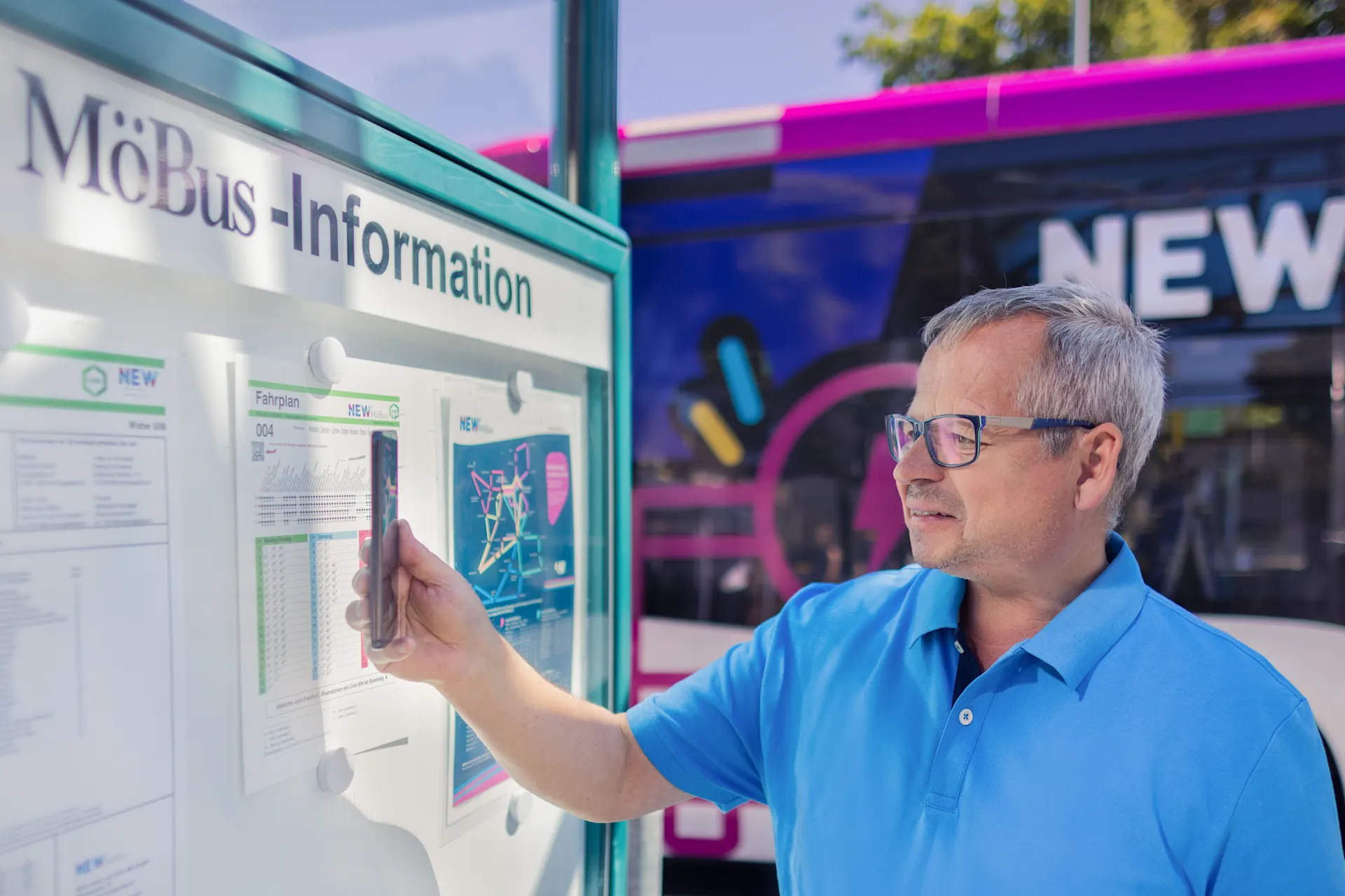 Ein Mann mit Brille und blauem Hemd steht vor einem weißen Informationsstand und zeigt auf einen Aushang. Im Hintergrund ist ein moderner Bus mit violetter und blauer Lackierung und der Aufschrift „NEW“ zu sehen.