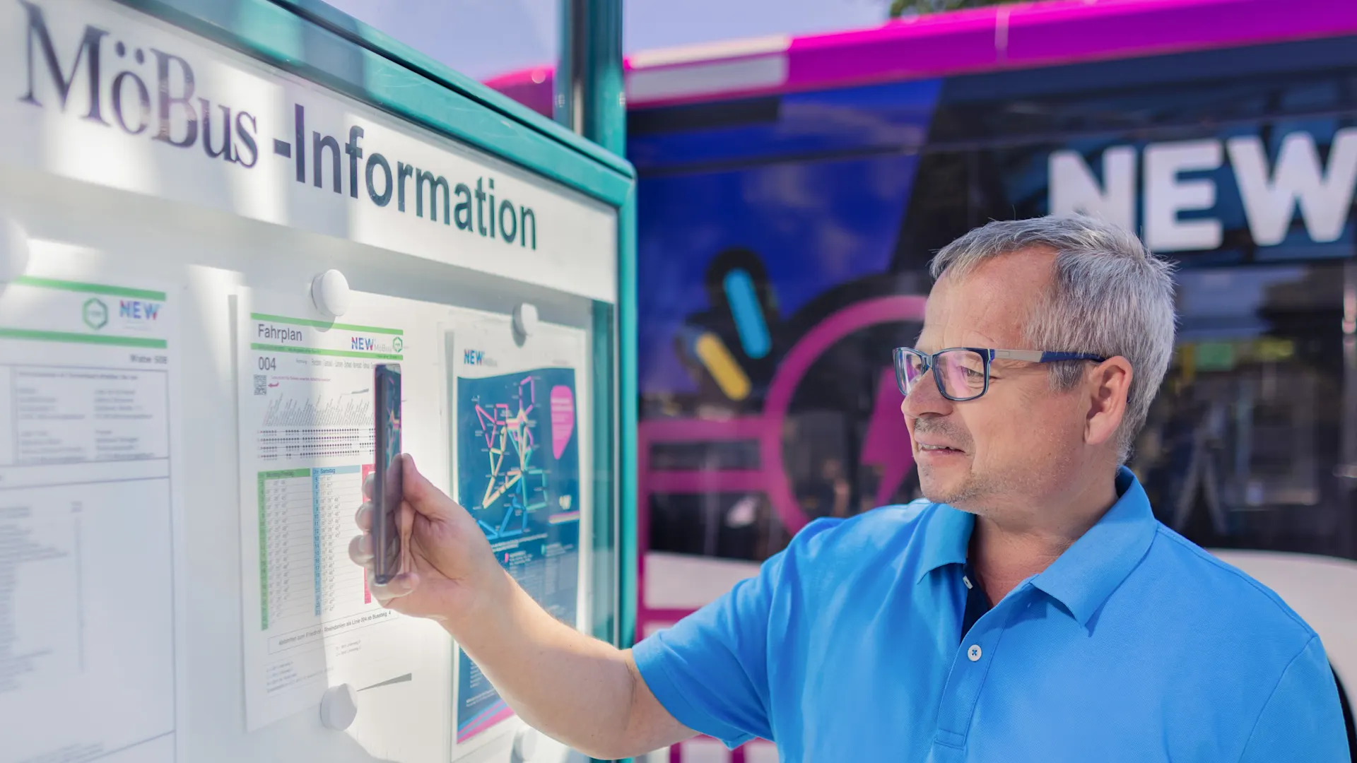 Clever unterwegs, nachhaltig voran. Ein Mann mit Brille und blauem Hemd steht vor einem weißen Informationsstand und zeigt auf einen Aushang. Im Hintergrund ist ein moderner Bus mit violetter und blauer Lackierung und der Aufschrift „NEW“ zu sehen.