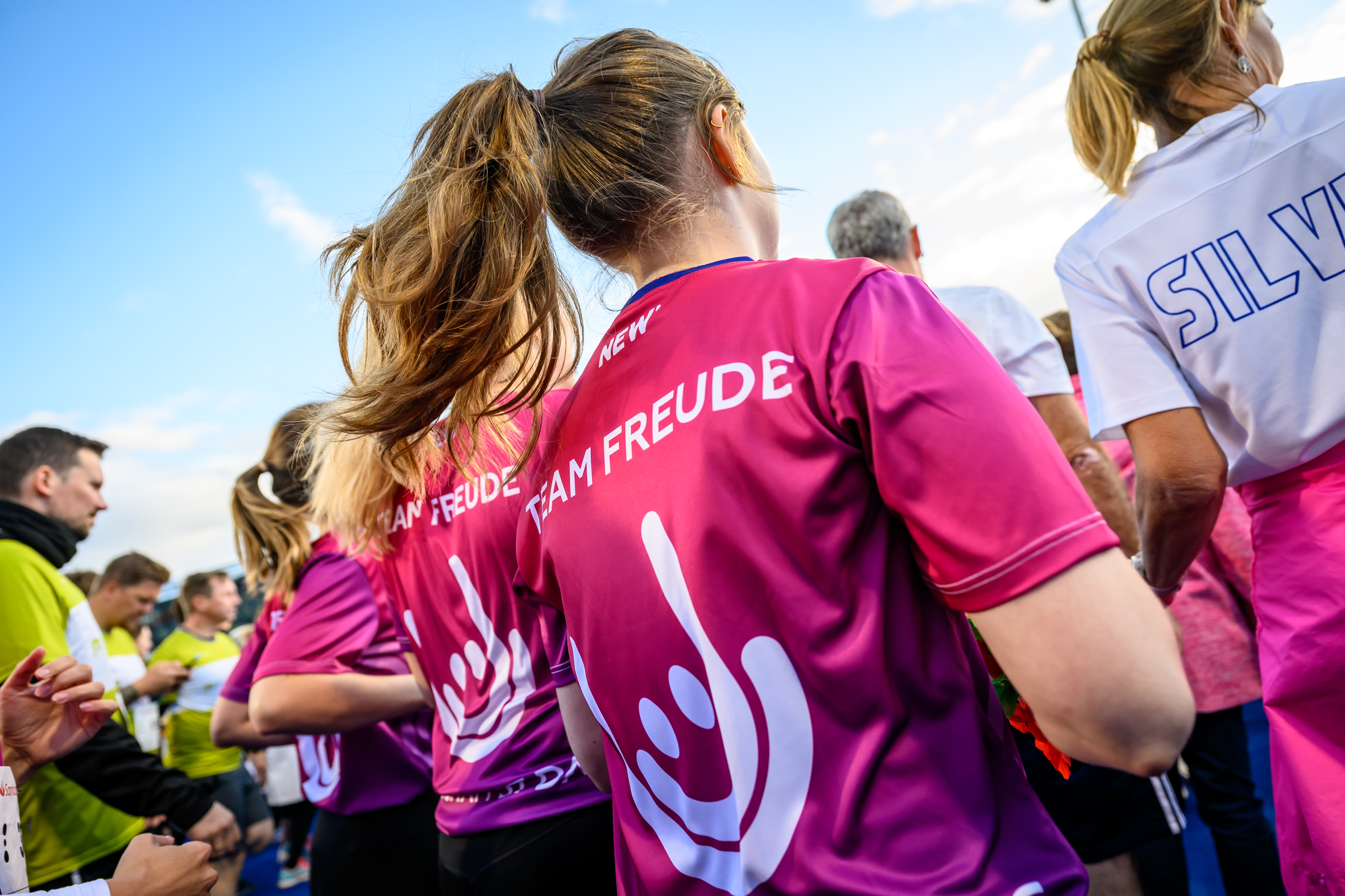 Rückenansicht von Läuferinnen in pinken Team-Shirts mit der Aufschrift „TEAM FREUDE“ bei einer Laufveranstaltung unter blauem Himmel. Die Teilnehmerinnen tragen Pferdeschwänze und stehen in einer größeren Gruppe von Läufer:innen, die sich zum Start eines Wettbewerbs versammelt haben. Im Hintergrund sind weitere Sportler:innen in gelben und weißen Trikots zu sehen.