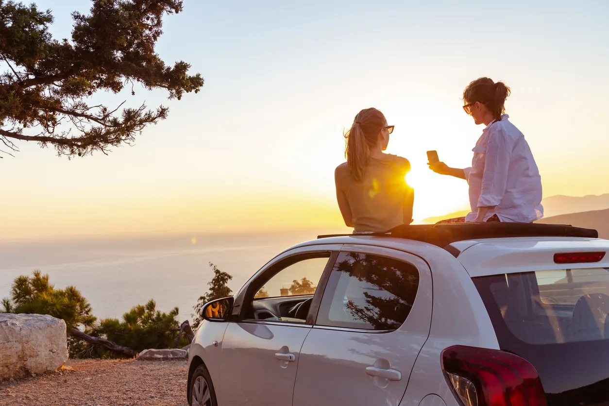 Zwei Personen sitzen bei Sonnenuntergang auf dem Dach eines weißen Autos. Eine Person hält ein Handy in der Hand und sie genießen die Aussicht auf die Küste bei Sonnenuntergang.