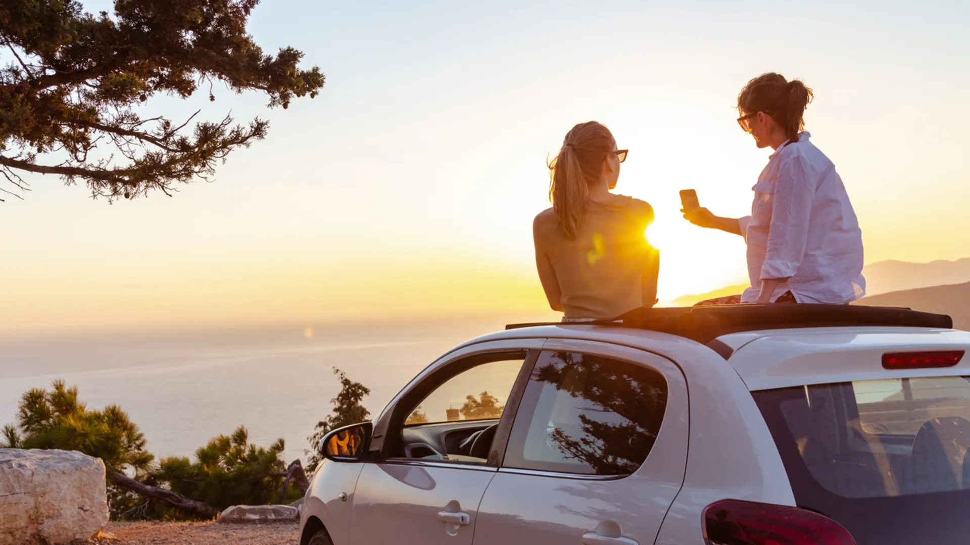 Urlaub mit E-Auto: stressfrei reisen mit Ladetipps, Routen & Zielen Zwei Personen sitzen bei Sonnenuntergang auf dem Dach eines weißen Autos. Eine Person hält ein Handy in der Hand und sie genießen die Aussicht auf die Küste bei Sonnenuntergang.